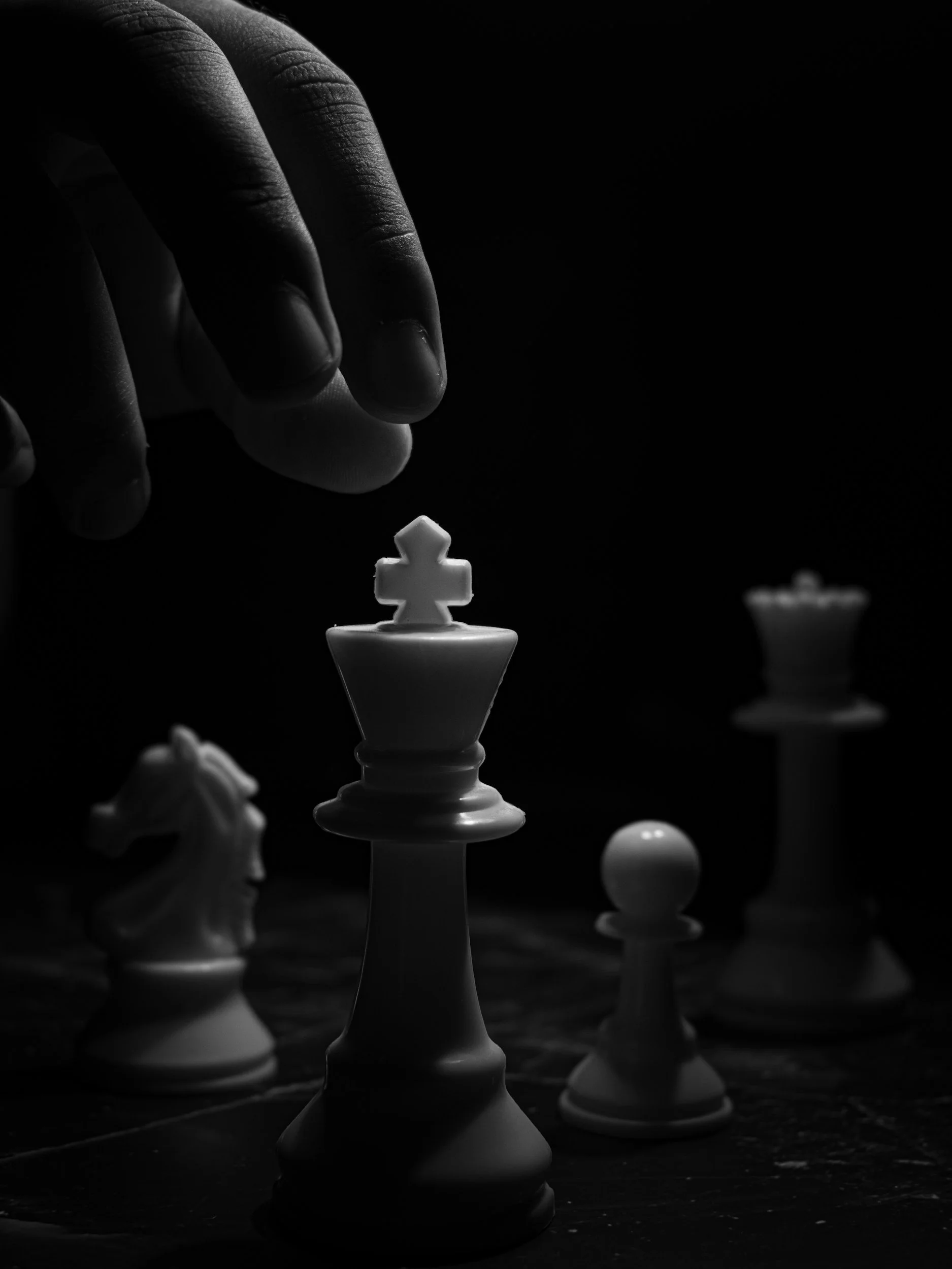 A black-and-white photograph of a chess game in progress with a black king piece in focus, hand moving a white pawn, and blurred chess pieces in the background.