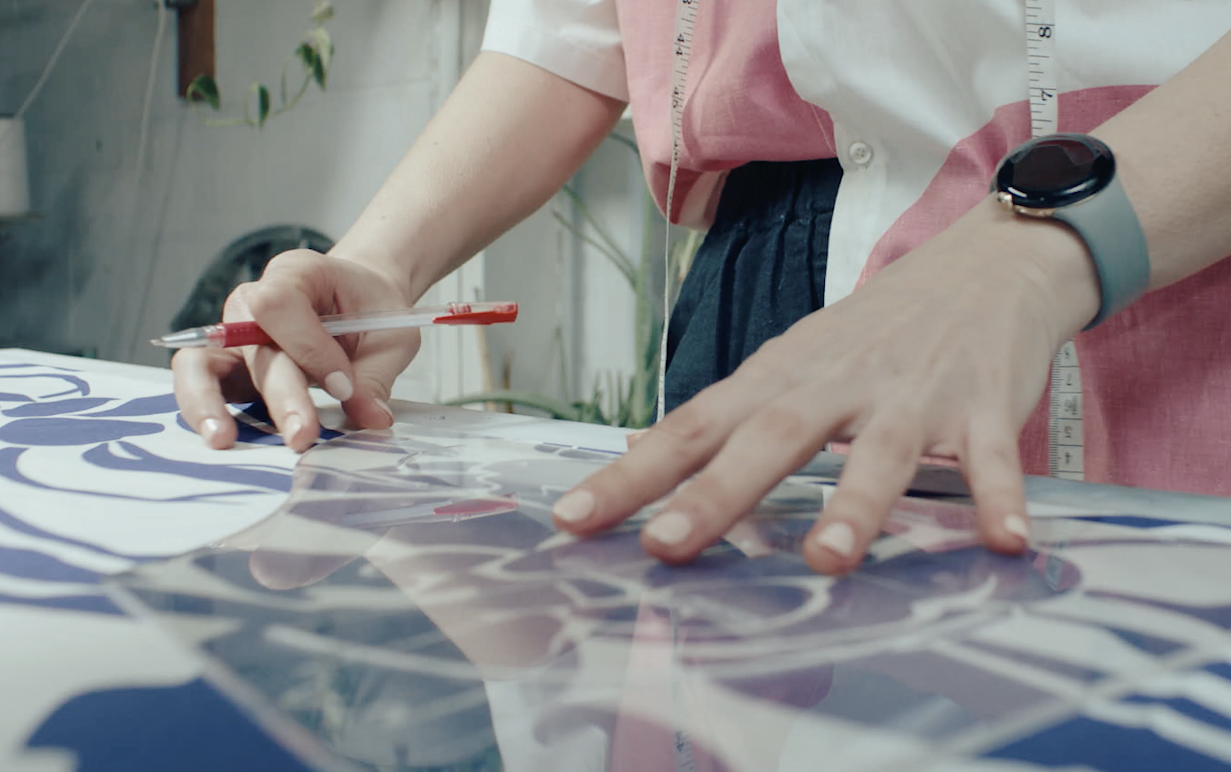 Close-up of a person working on a craft project, measuring and cutting patterned paper on a table, wearing a watch and pink shirt with a measuring tape.