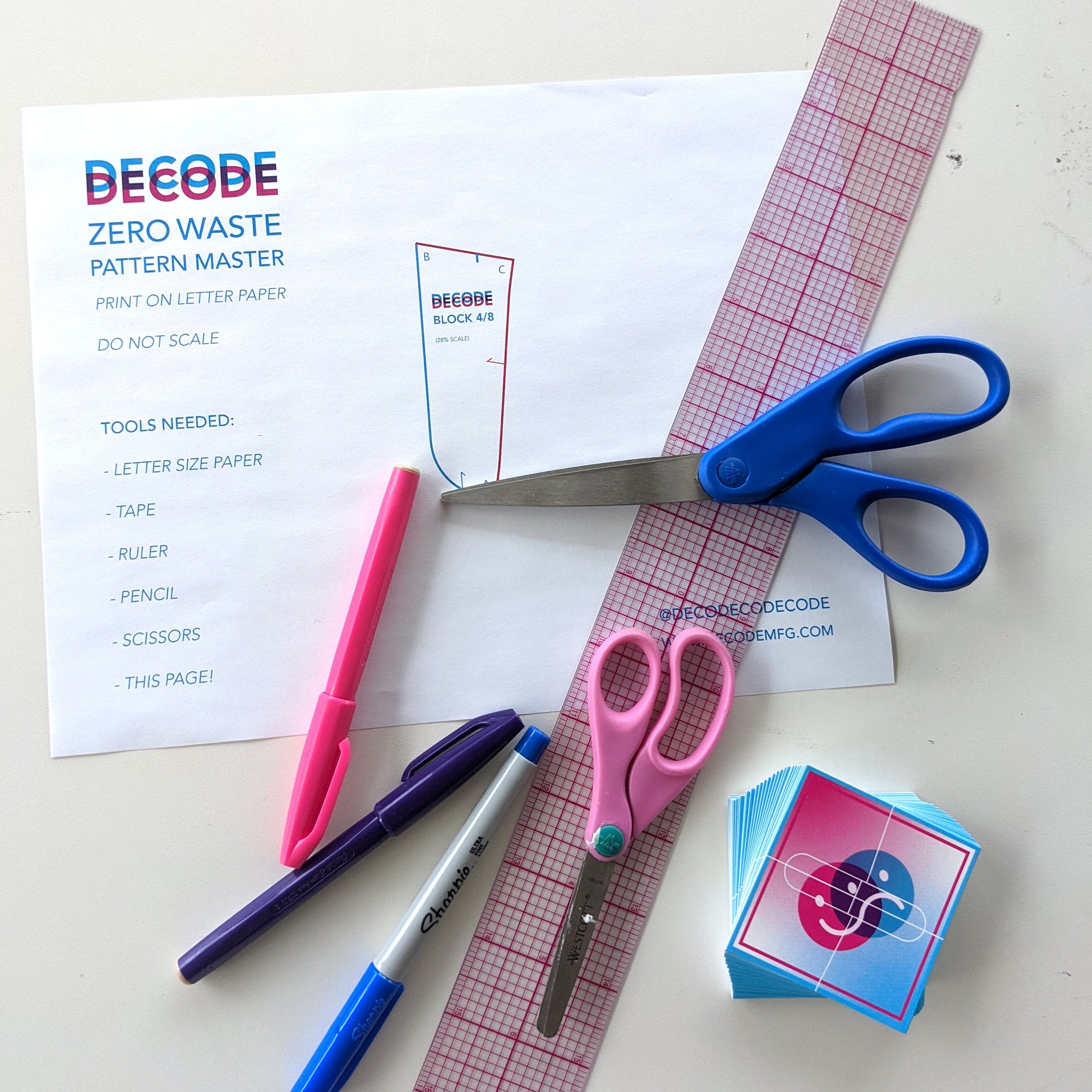 A collection of stationery items including a pair of blue scissors, pink and blue markers, a silver ruler, pink cutting scissors, a pink marker, a pink grid cutting mat, a stack of colored stickers, and a printed pattern master sheet titled 'Decode Zero Waste Pattern Master' with various tools needed listed.