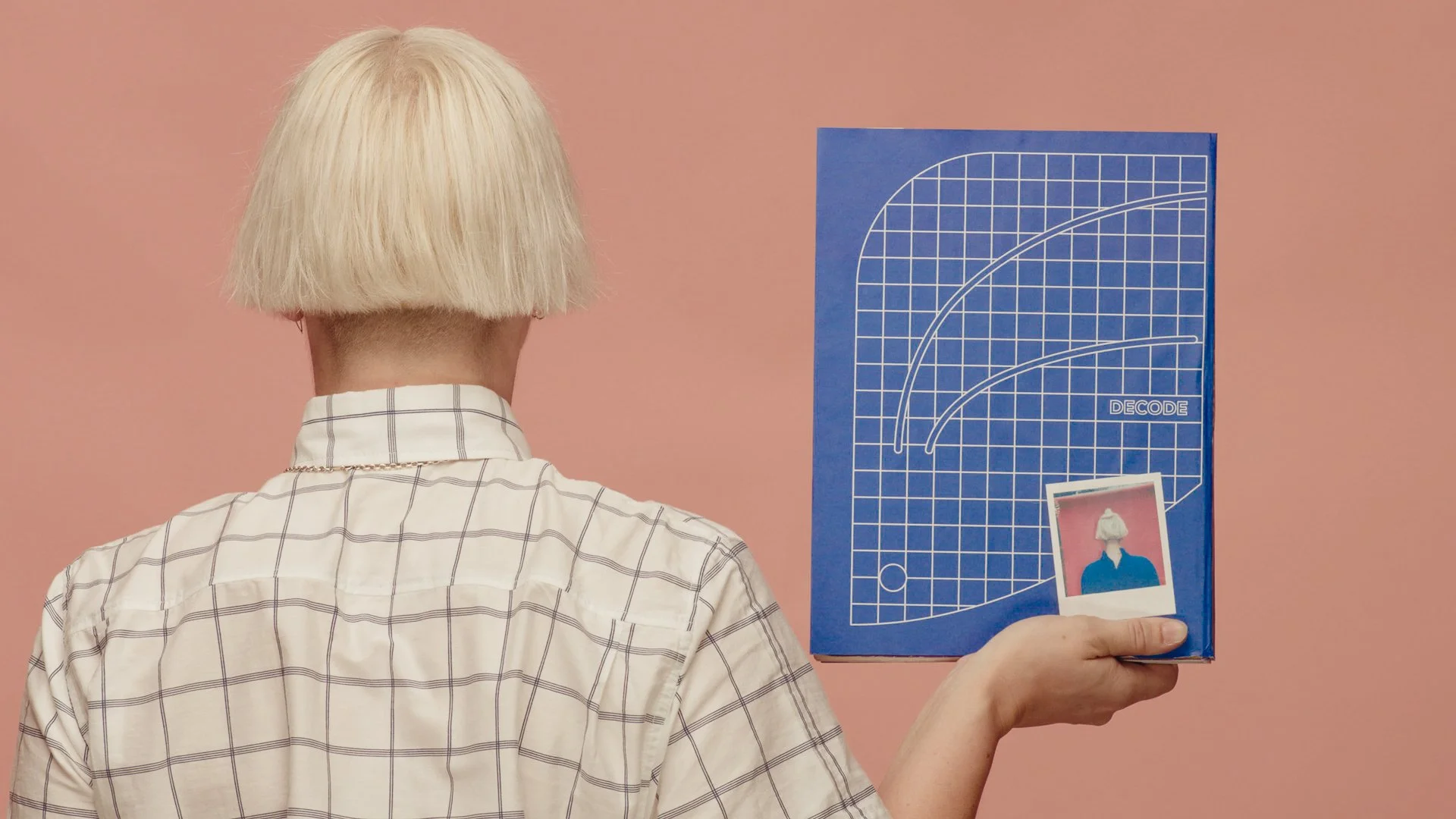 Person with blond hair in a beige checkered shirt holding a blue poster with curved white lines and the word 'DECODE,' in front of a pink wall. They are also holding a Polaroid photograph of a person with blond hair in front of a pink background.