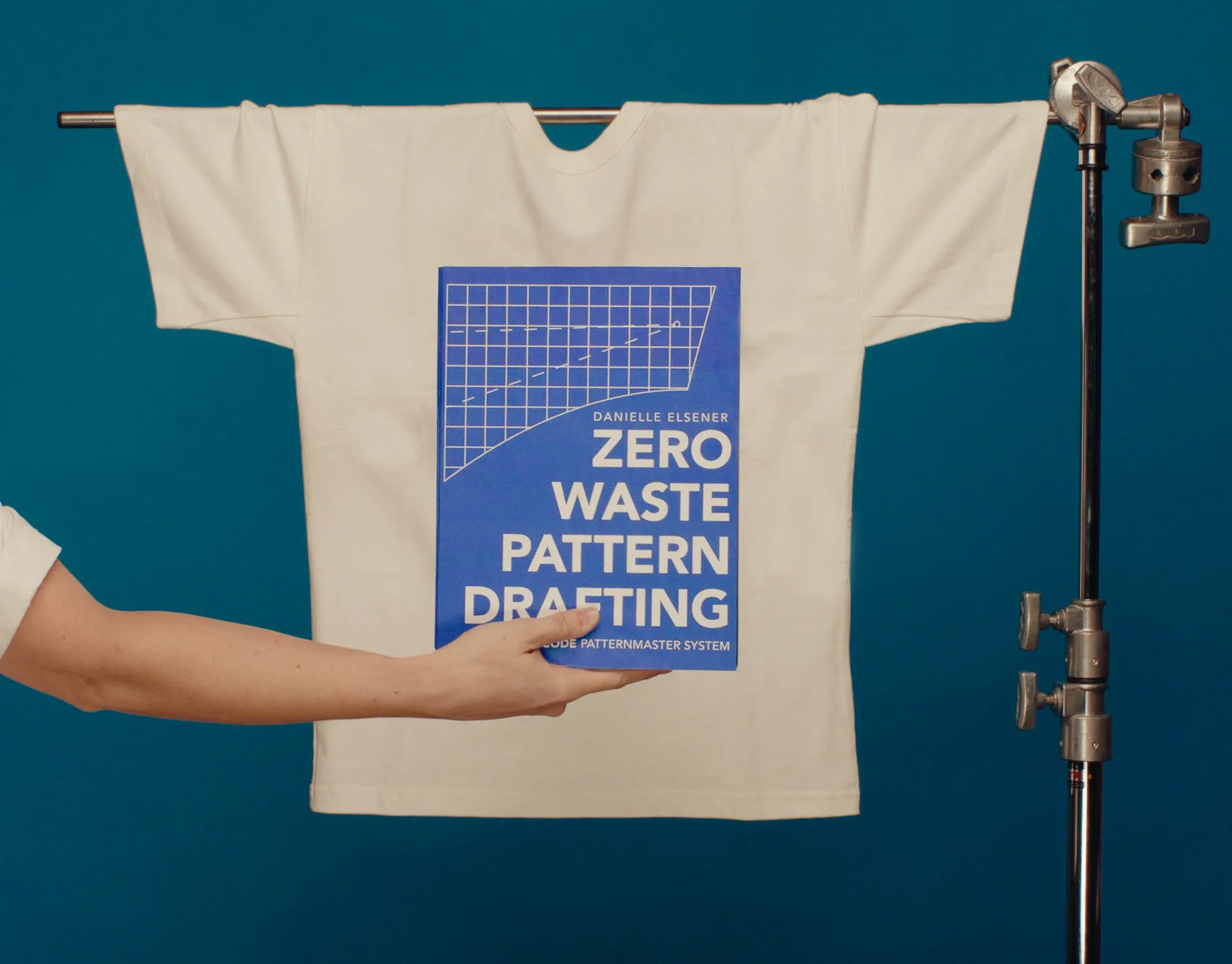 White T-shirt hanging on a clothing rack, with a blue pattern drafting book titled 'Zero Waste Pattern Drafting' held by a person. The background is blue.