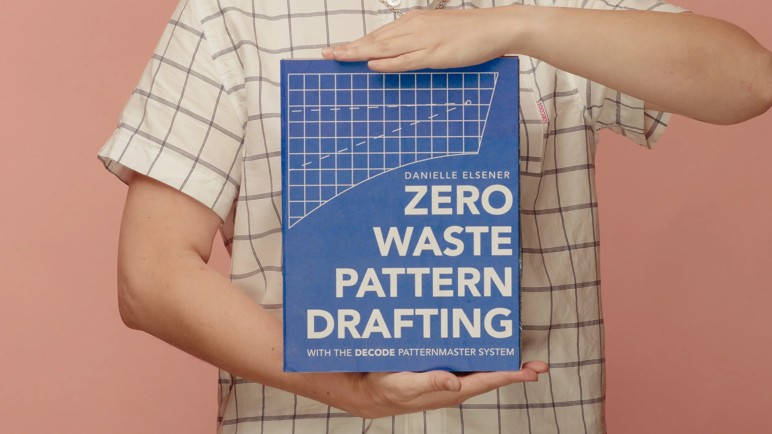 Person holding a blue book titled "Zero Waste Pattern Drafting" by Danielle Elsener, with a chart graphic on the cover, against a pink background.