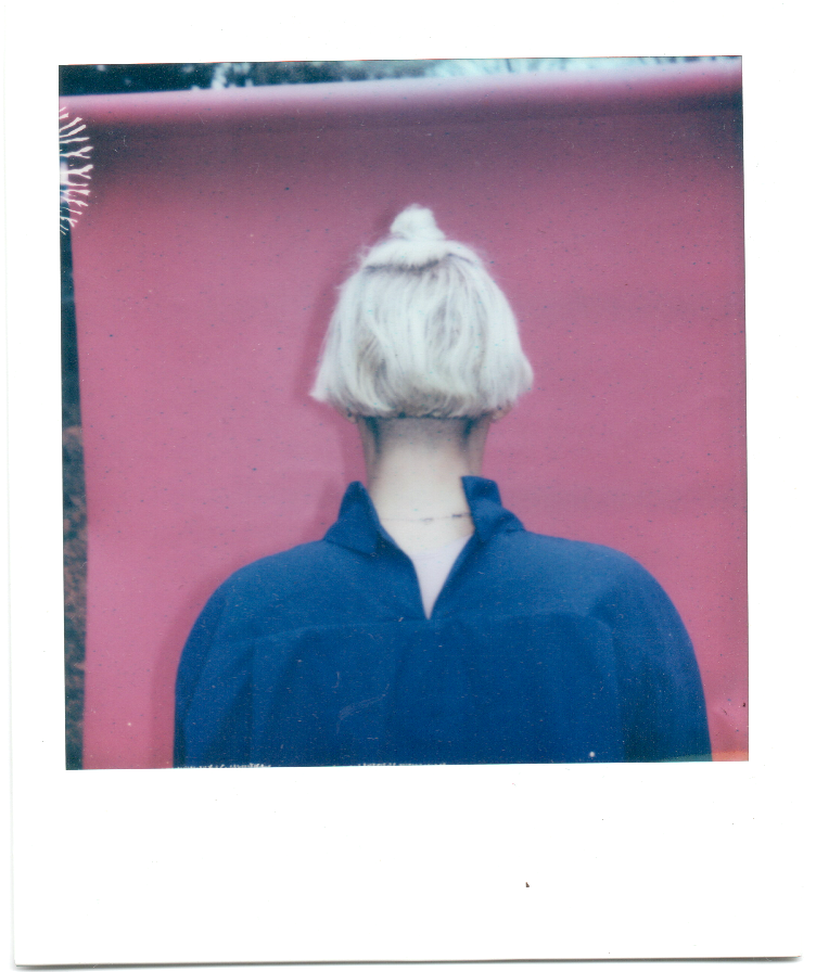 A person with short blonde hair wearing a dark blue hoodie facing a pink wall.