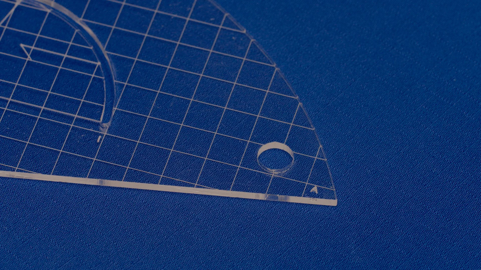 A clear acrylic ruler with grid lines, a rounded cutout, and an A marking, placed on a blue textured surface.