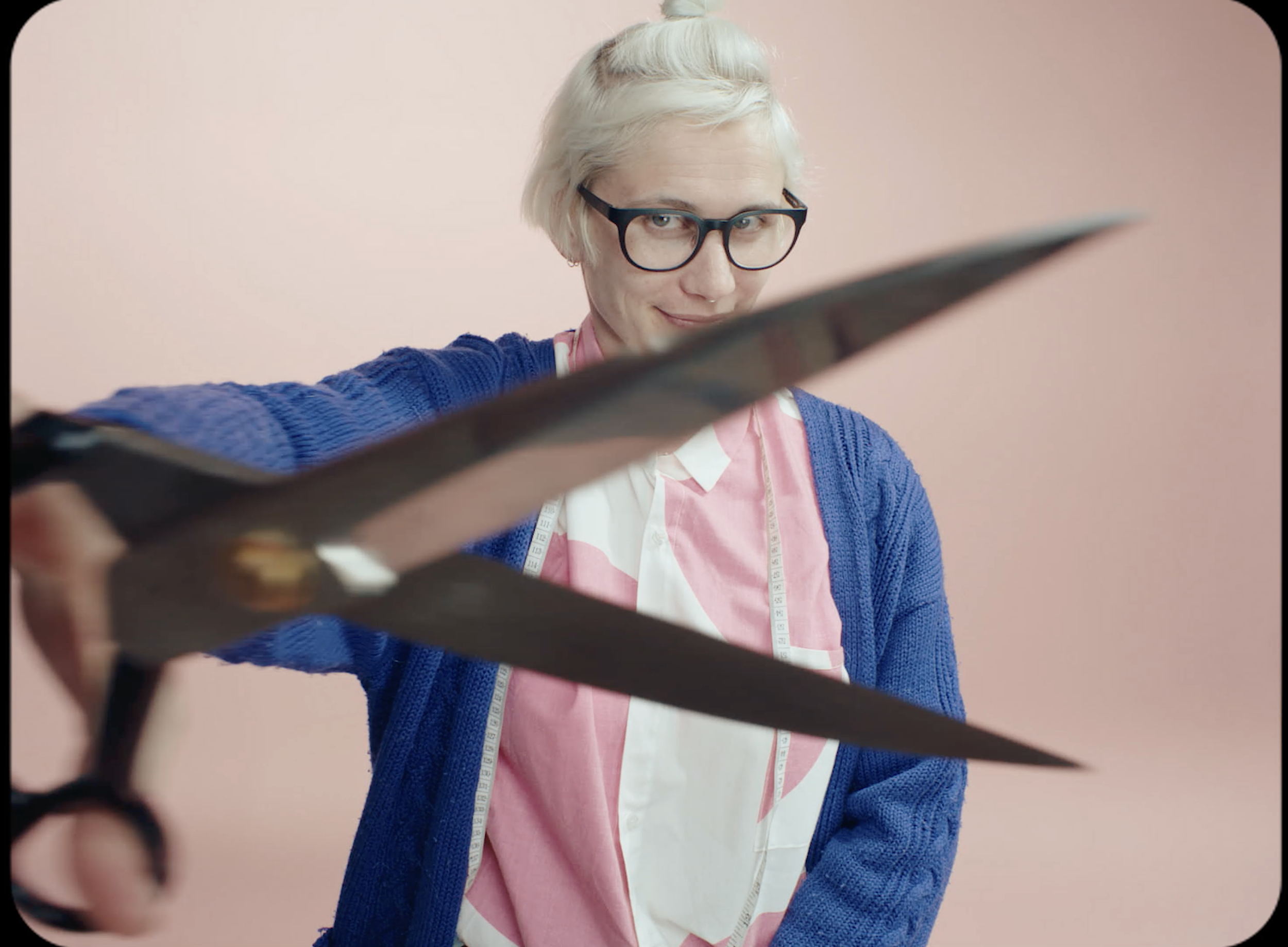 A person with short, white hair and glasses smiling while holding a scissors. They are wearing a pink and white apron over a white shirt and a blue knitted sweater, standing against a pink background.