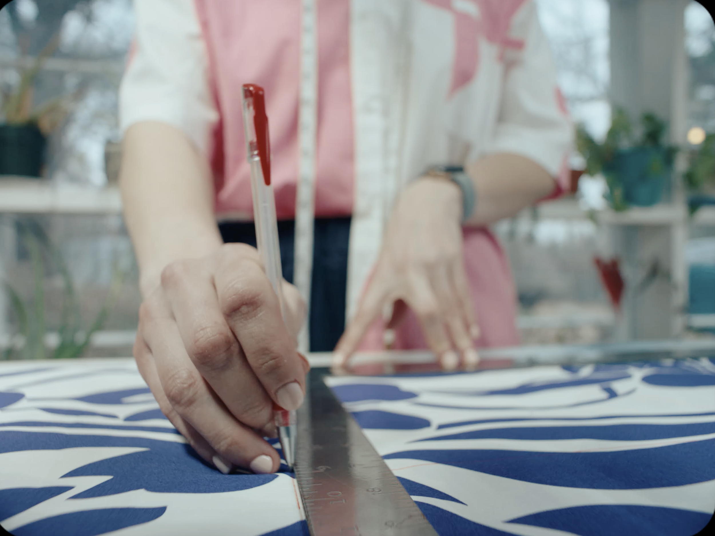 A person's hand is measuring fabric with a metal ruler in a sewing or craft room. The fabric has a blue and white pattern. Shelves with plants and other items are visible in the background.
