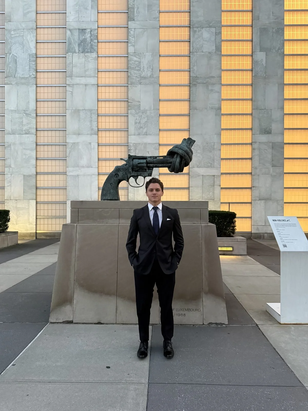 Lucas A. Perry in a black suit and blue tie beside the Knotted Gun sculpture at United Nations Headquarters in New York City.