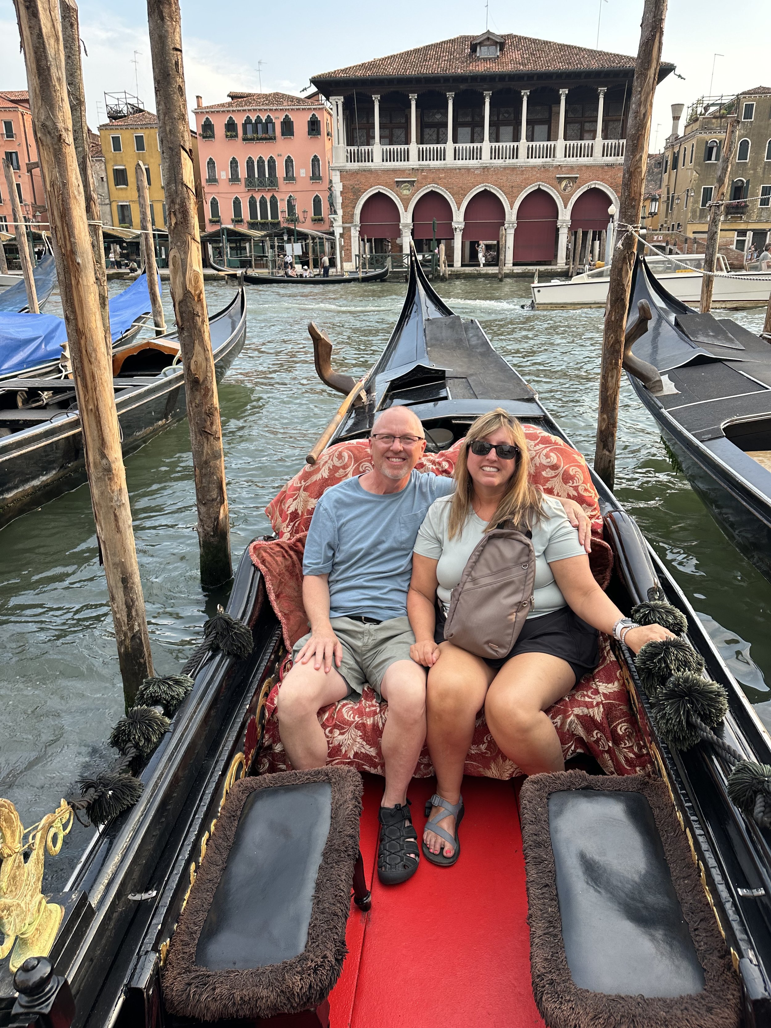 Couple sitting in a gondola on a canal in Venice, Italy, with colorful buildings and other gondolas visible in the background.