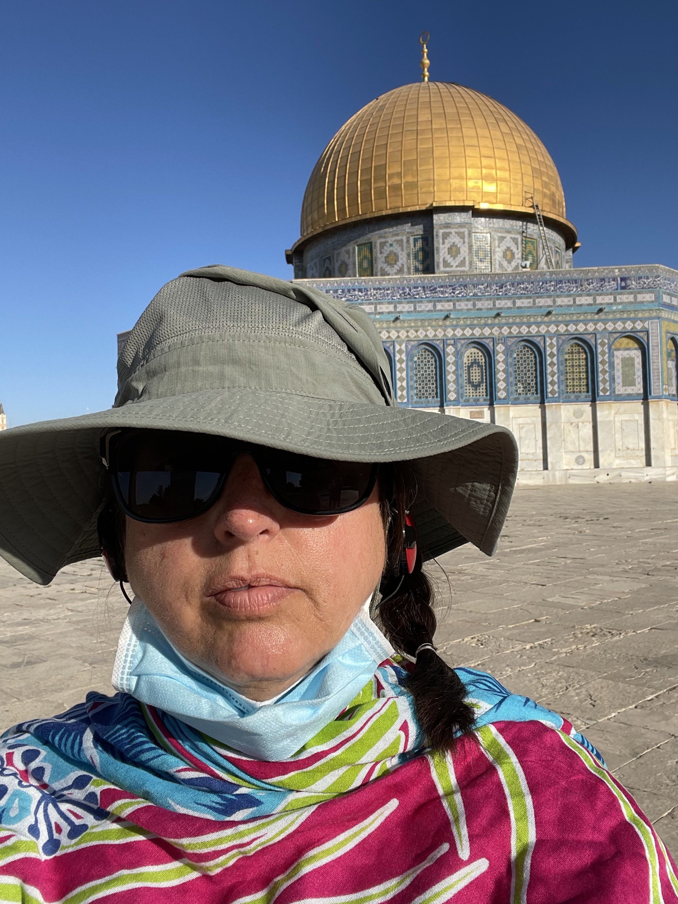 Close-up of a woman wearing a large sun hat, sunglasses, a face mask pulled down, and colorful clothing, standing in front of the Dome of the Rock with its golden dome and intricate mosaic exterior under clear blue sky.