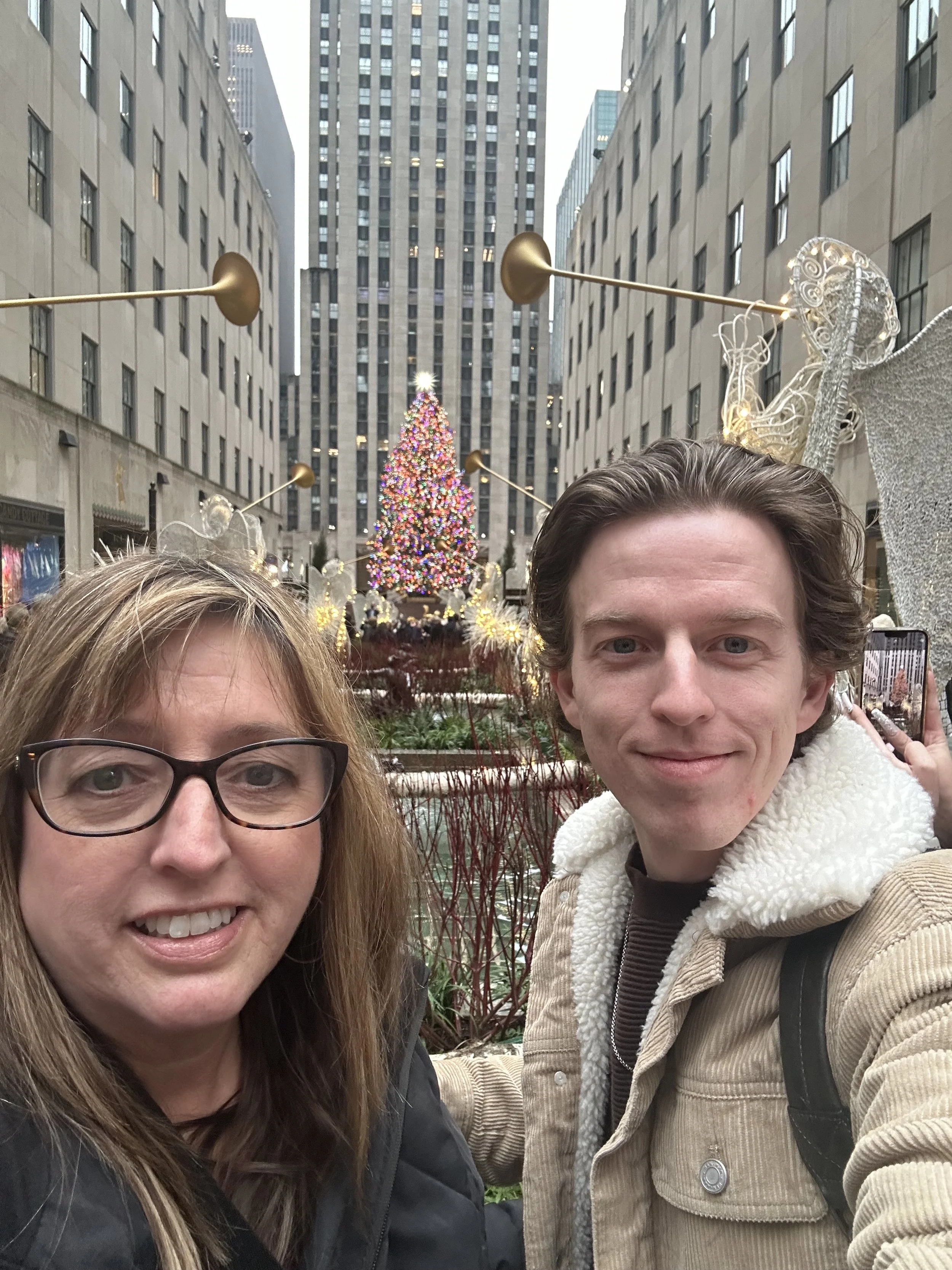 A woman with glasses and a young man smiling for a selfie in front of a decorated Christmas tree in an urban setting with tall buildings.