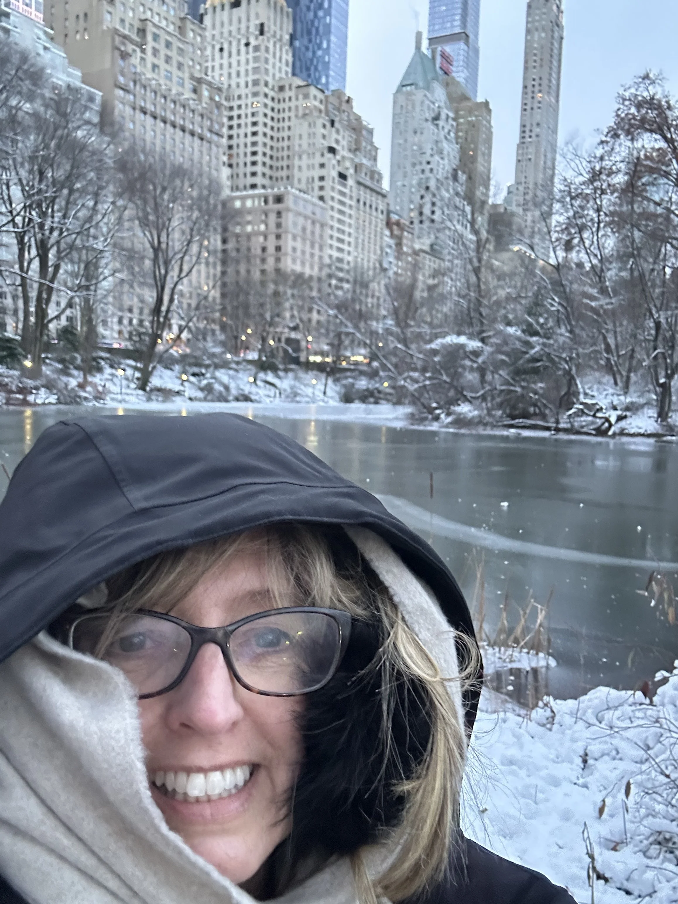 A woman wearing glasses and a winter coat smiling in front of a snowy park with trees and tall buildings in the background.