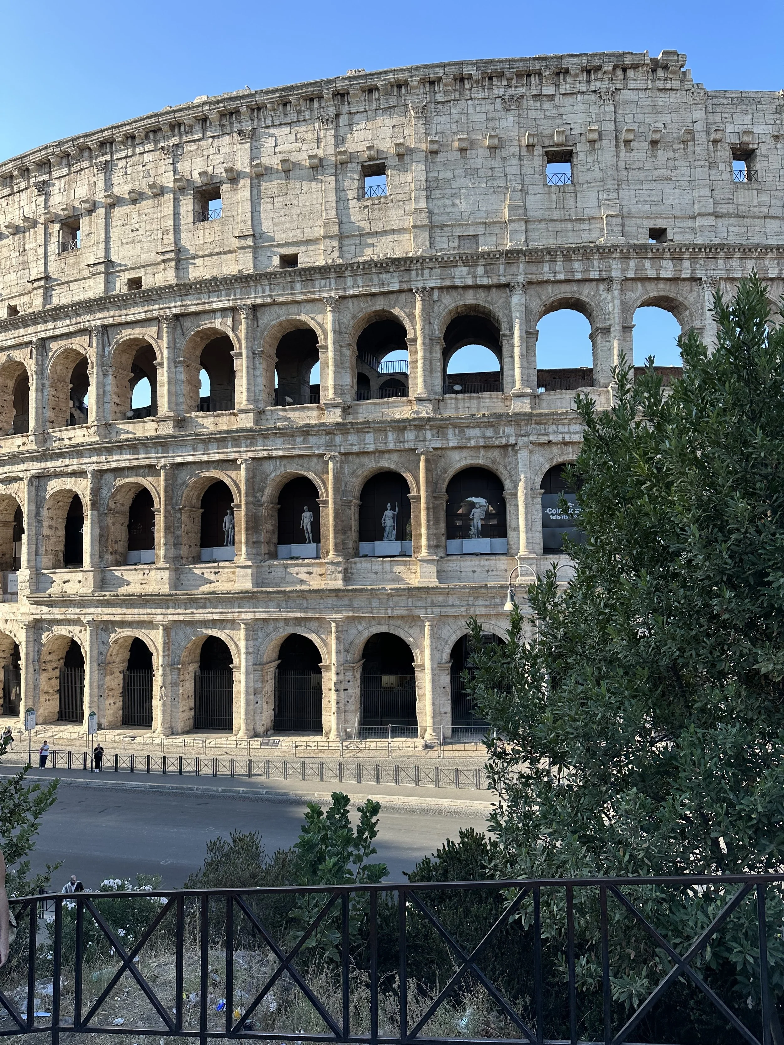 A view of the Colosseum in Rome, Italy, showing its large stone arches and partial upper levels, with some small windows and statues, under a clear blue sky.