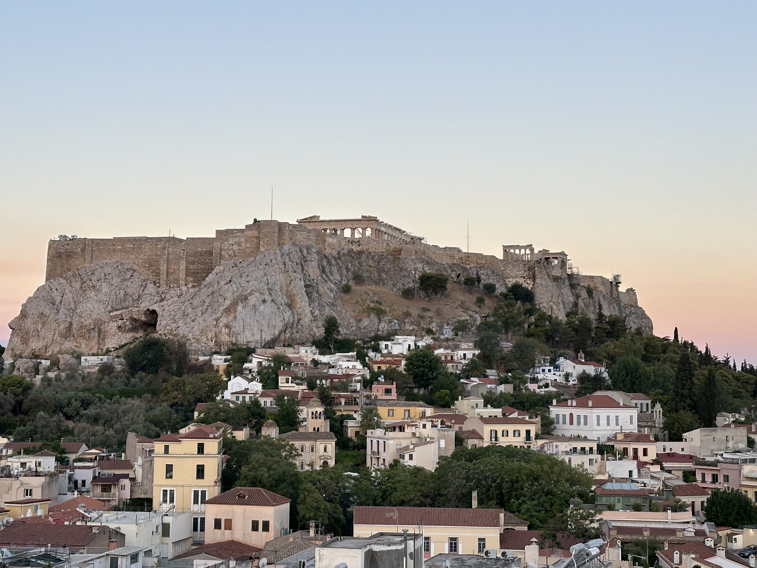 The Acropolis of Athens with ancient ruins on a hilltop, overlooking a city with residential buildings and lush greenery at sunset.