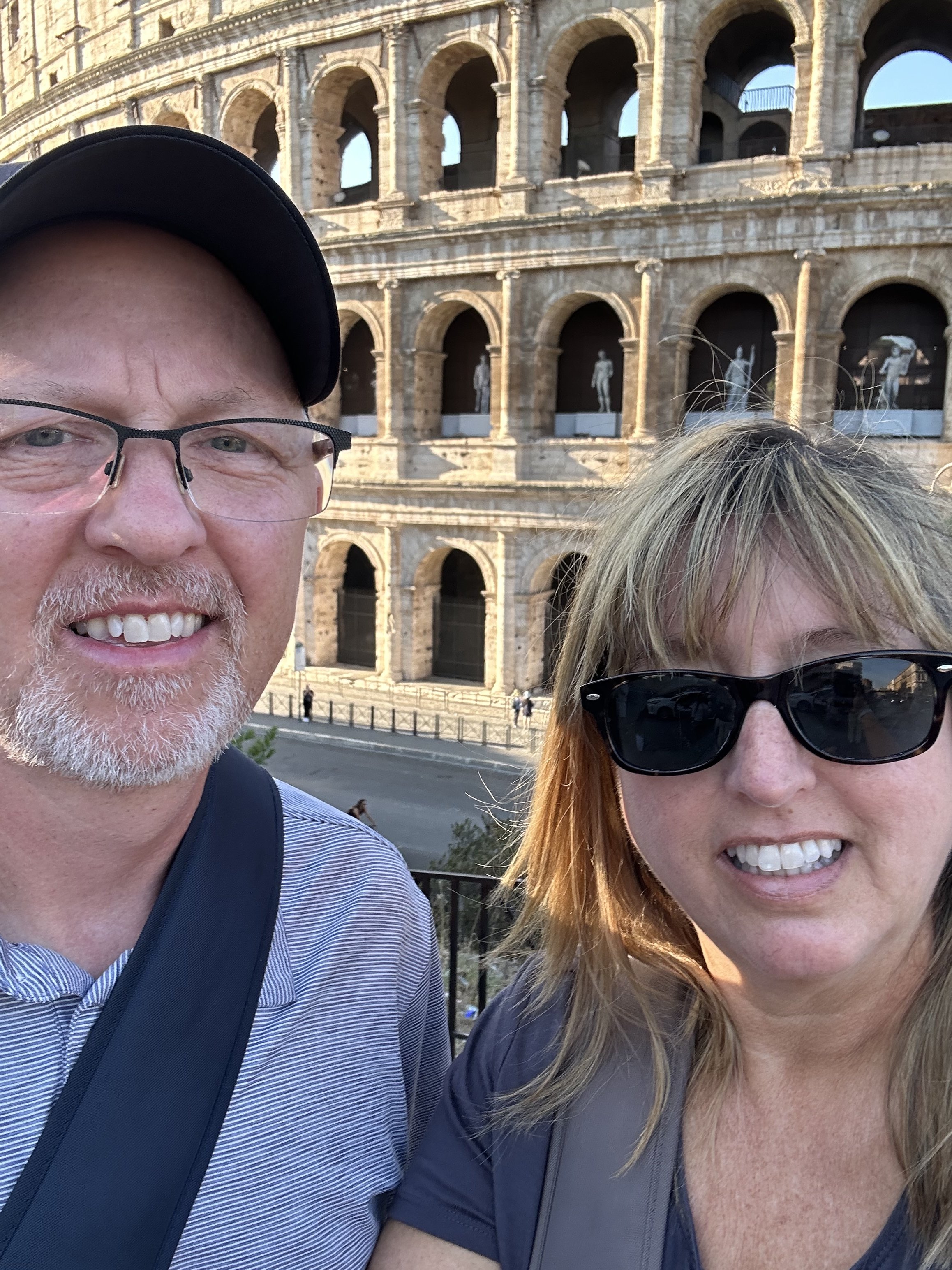A selfie of a man and a woman smiling in front of the Roman Colosseum in Rome, Italy, during daytime.