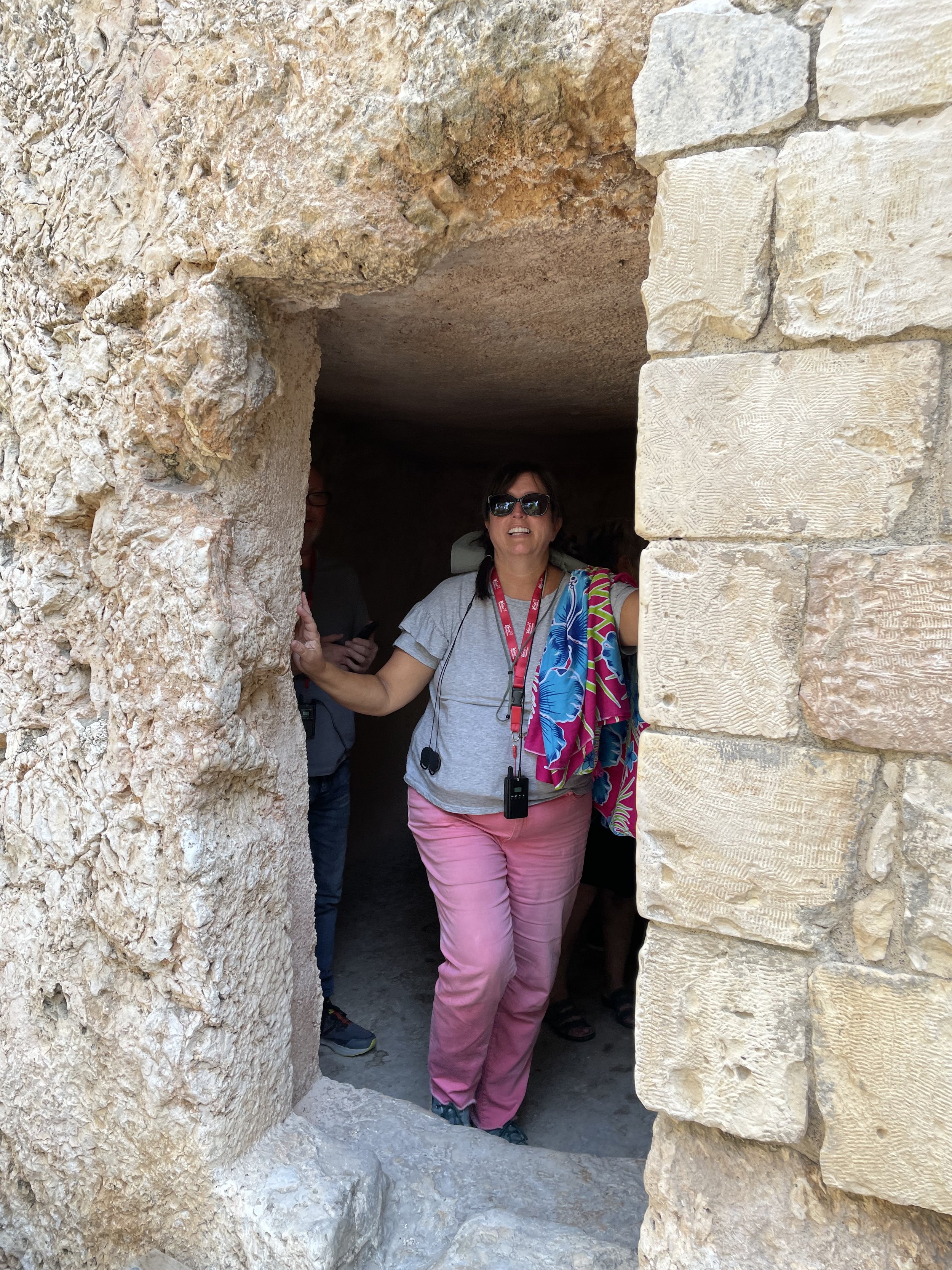 A woman wearing sunglasses and pink pants is standing at an ancient stone doorway of Jesus's burial site with her hand on the rough stone wall. She has a colorful scarf draped over her shoulder.