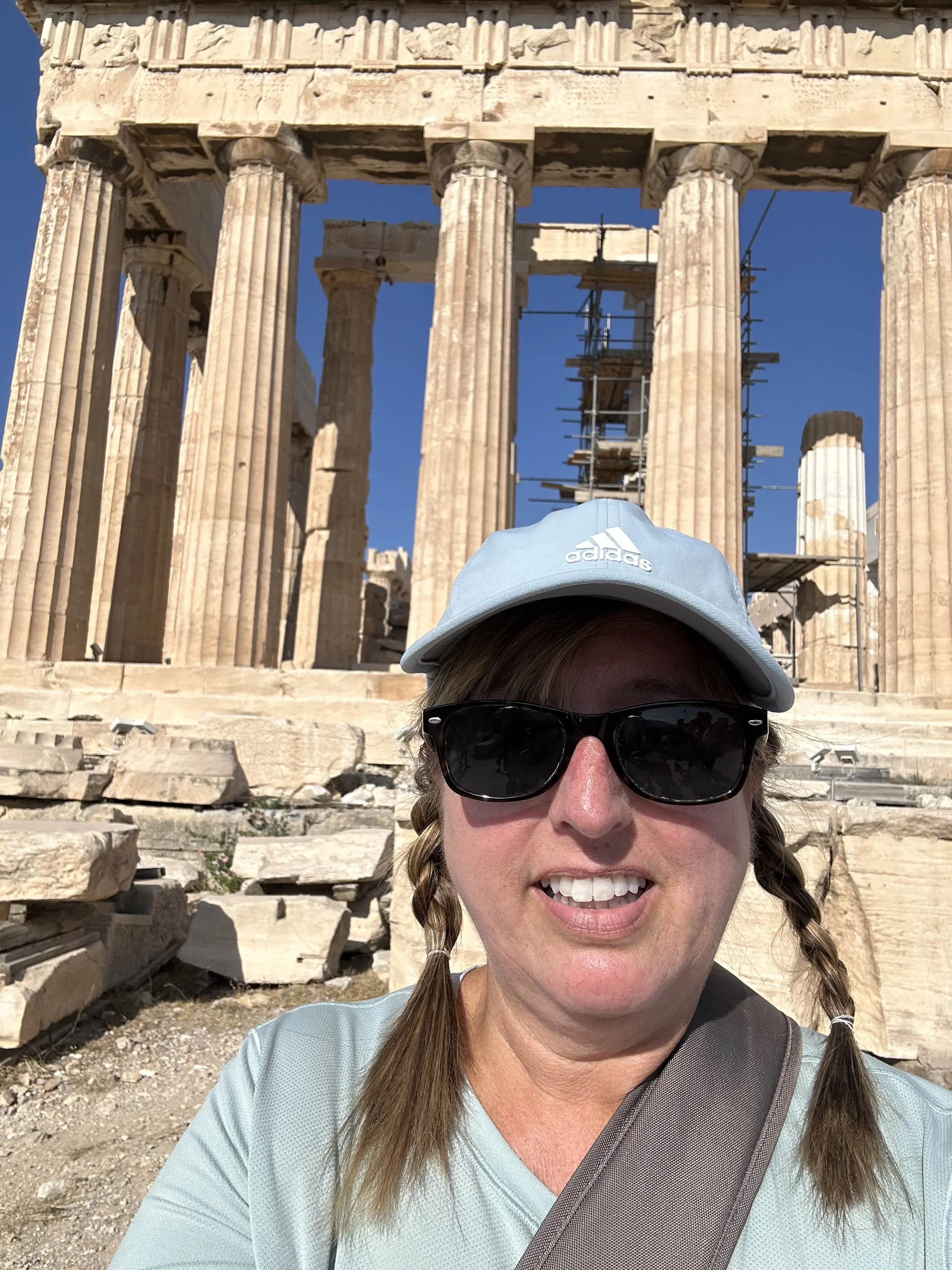A woman wearing a light blue cap, sunglasses, and a blue shirt in front of ancient Greek ruins with columns and scaffolding.