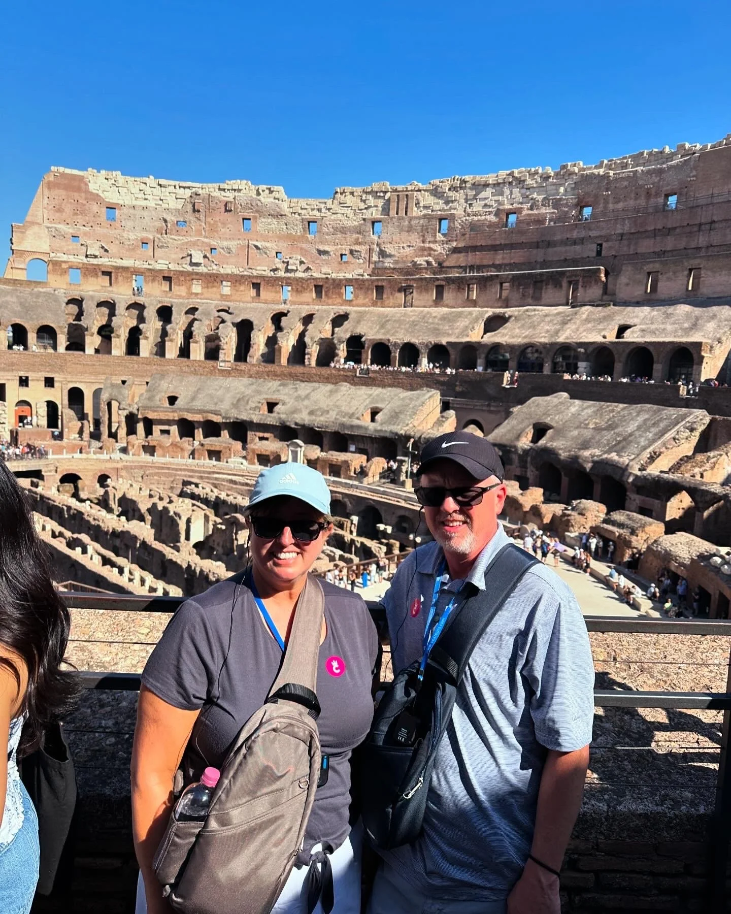 Two smiling tourists standing inside the Colosseum in Rome, Italy, with the ancient amphitheater's arches and tiers in the background against a clear blue sky.