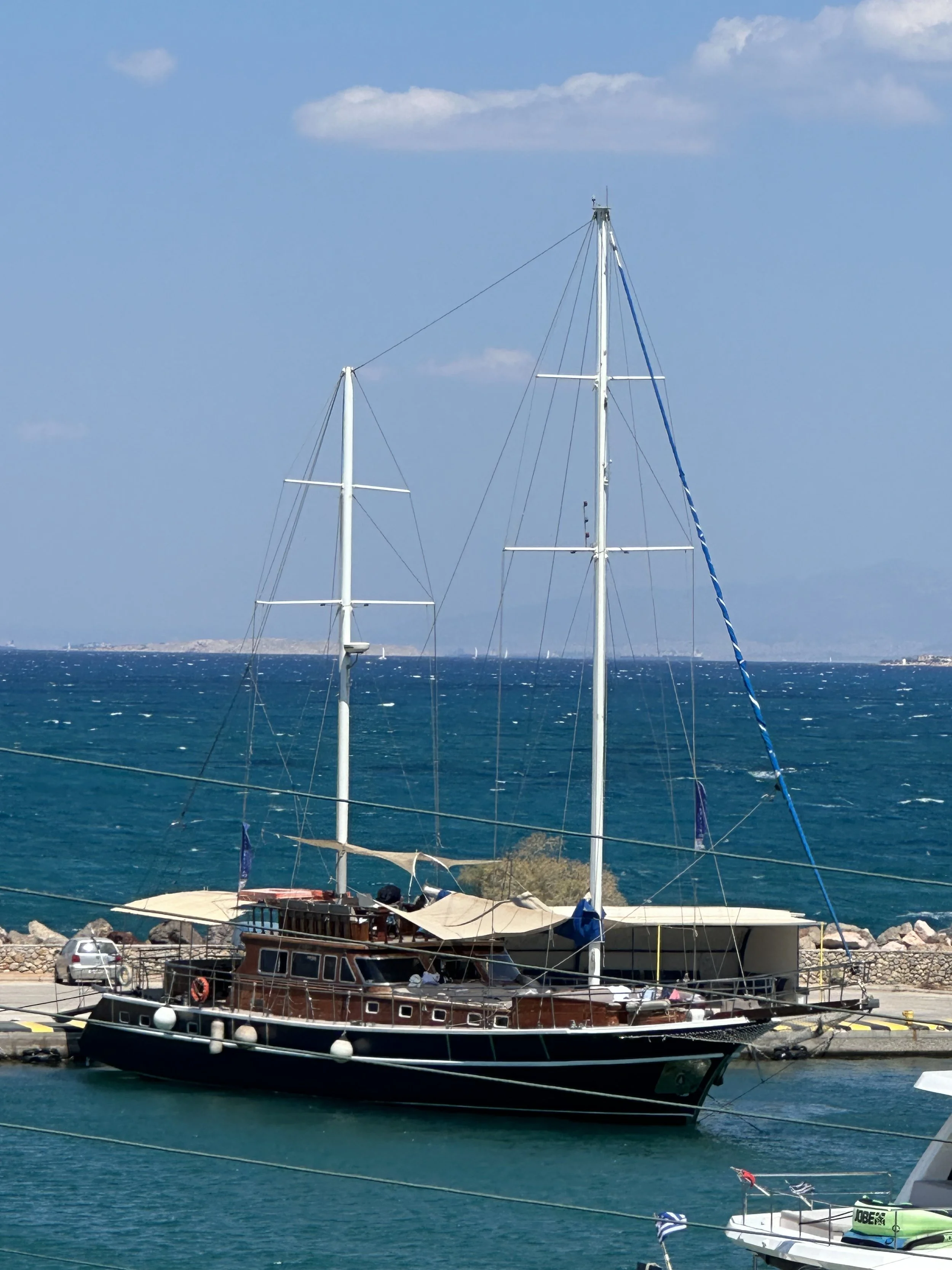 Large sailboat docked at a marina with the sea and a coastline in the background. The sailboat has tall masts and a wooden hull, with some other smaller boats around.