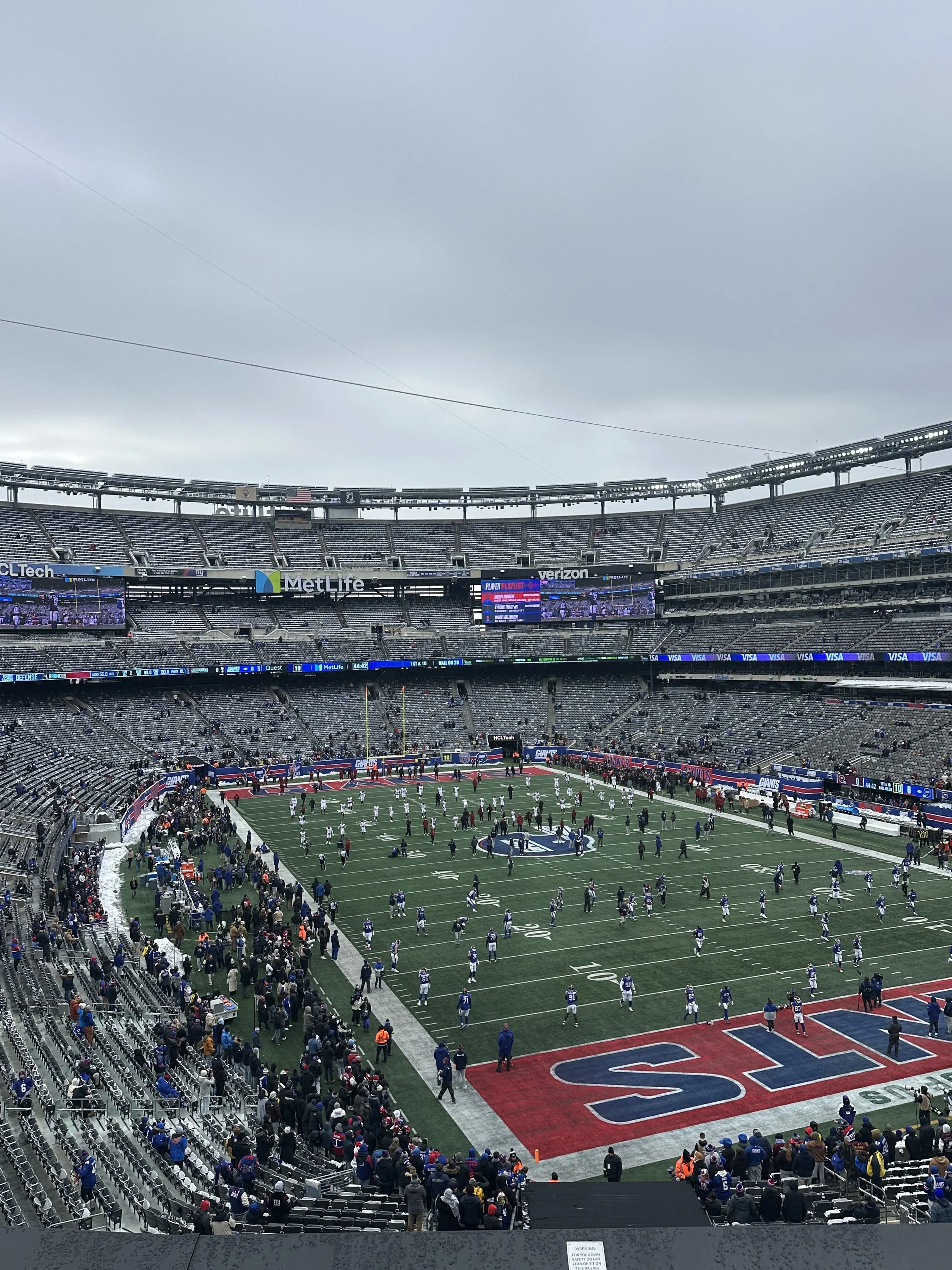 An outdoor football stadium with fans seated in the stands, players on the field, and a cloudy sky overhead. The scoreboard displays a game in progress.
