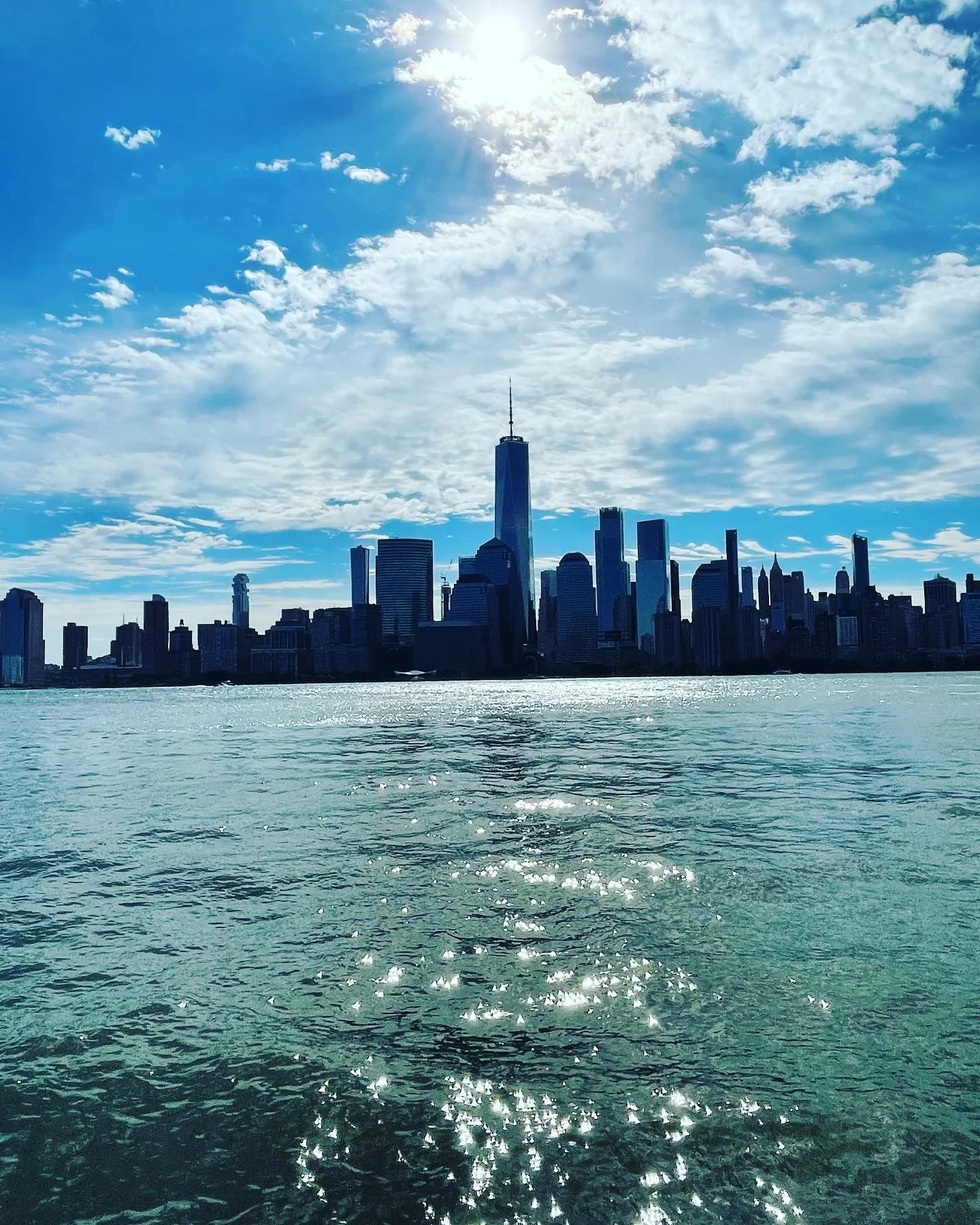 New York City skyline with One World Trade Center, viewed from the water under partly cloudy sky.
