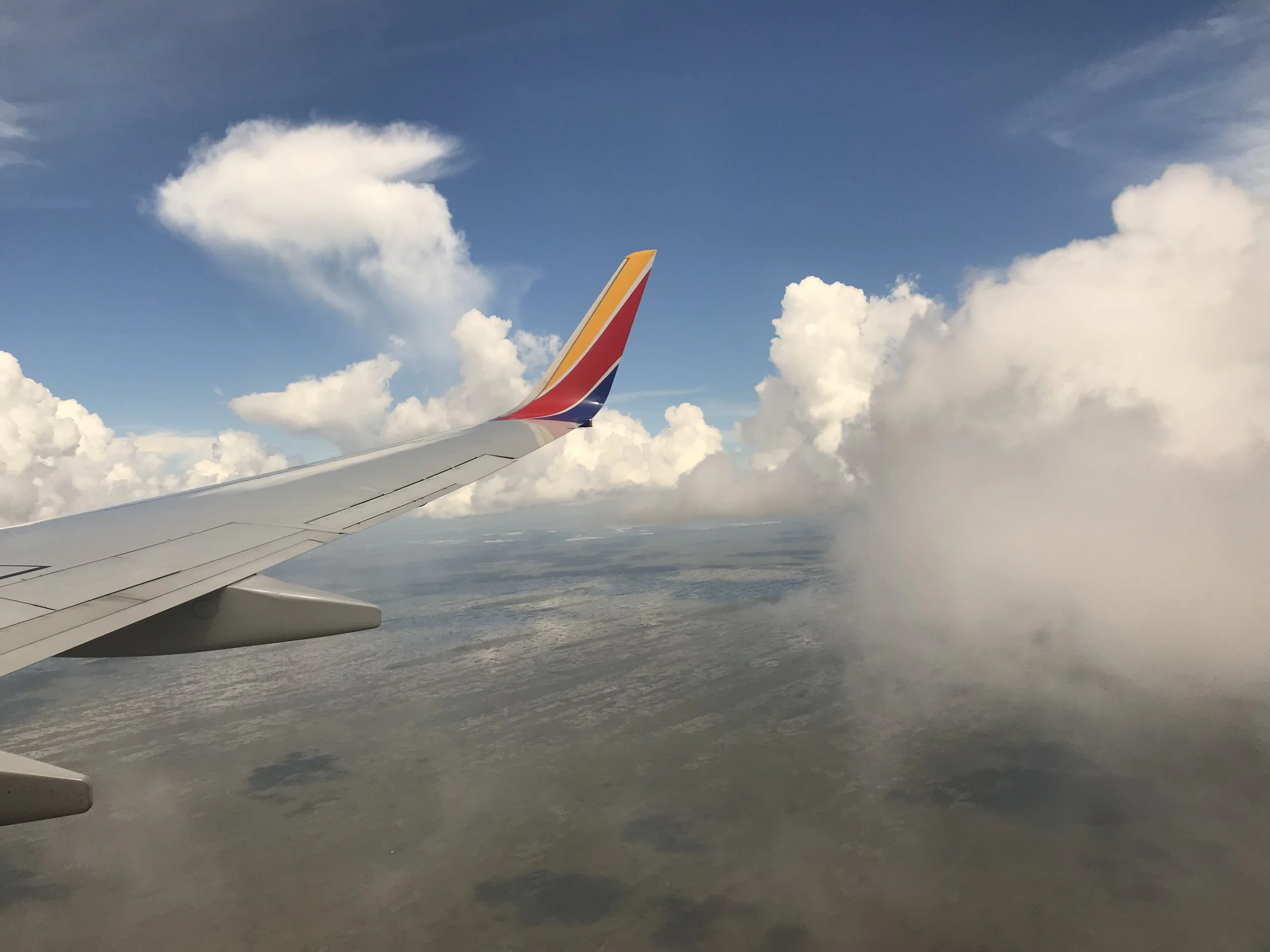 View from an airplane window showing the airplane wing with the airline logo on the wingtip against a sky filled with clouds and a landscape below.
