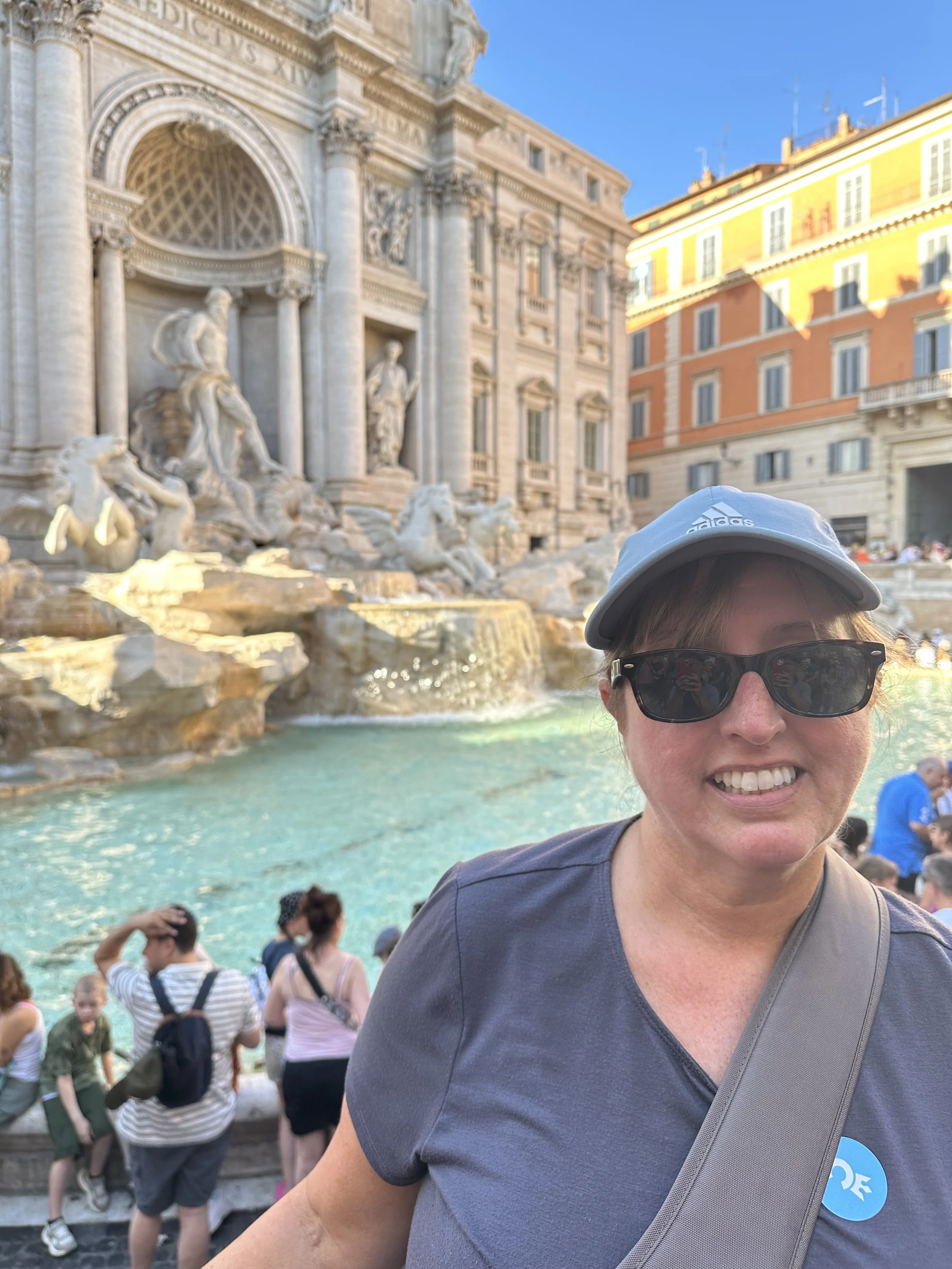 A person in a gray t-shirt, sunglasses, white cap, and backpack taking a selfie in front of the Trevi Fountain in Rome, Italy, with tourists and historic buildings in the background.