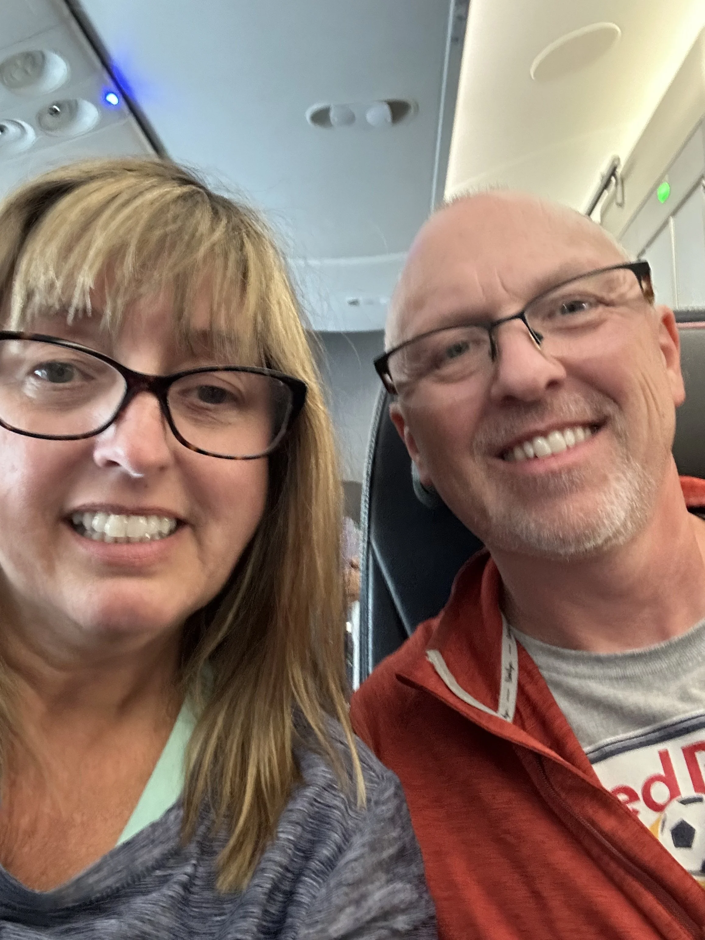 A selfie of a woman and a man sitting next to each other on a plane. The woman has blonde hair, glasses, and is smiling. The man is bald, has glasses, and is smiling. The airplane interior, including overhead compartments, can be seen in the background.