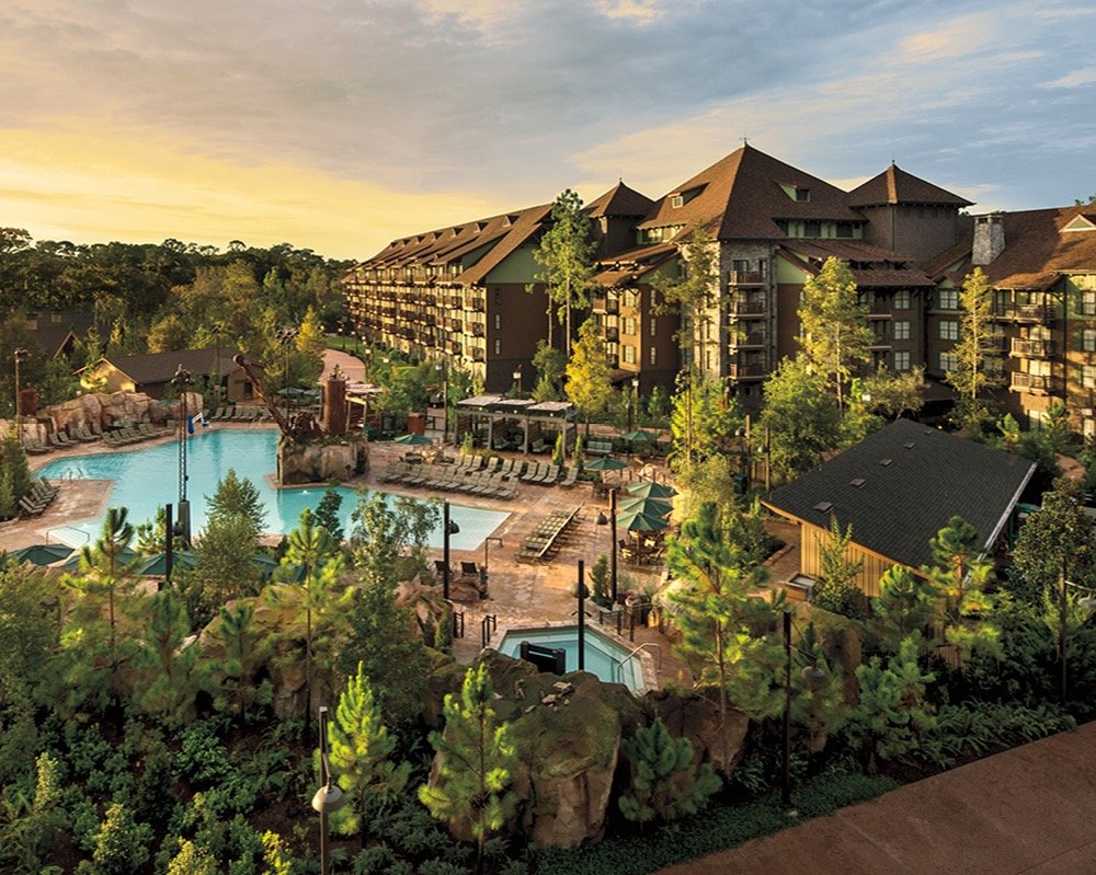 A large resort-style complex with a main building, a pool area, smaller pools, lounge chairs, umbrellas, and surrounding trees and greenery during sunset.