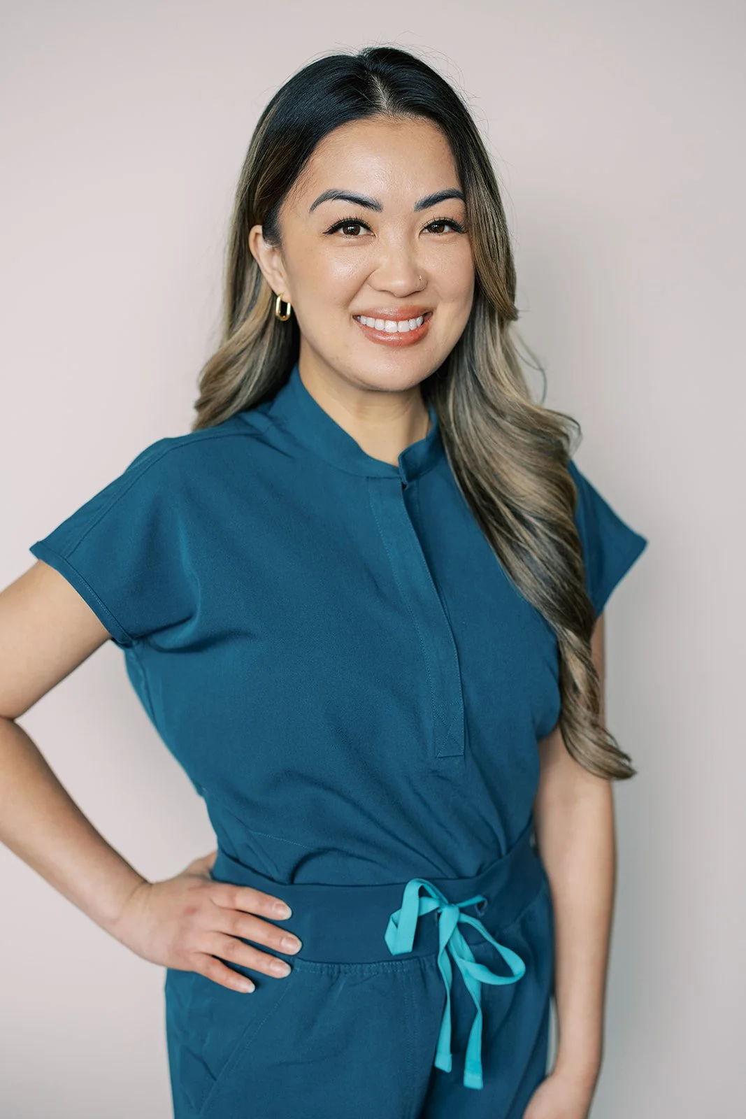 A woman with long hair, wearing a blue medical uniform, smiling with her hand on her hip against a plain light background.