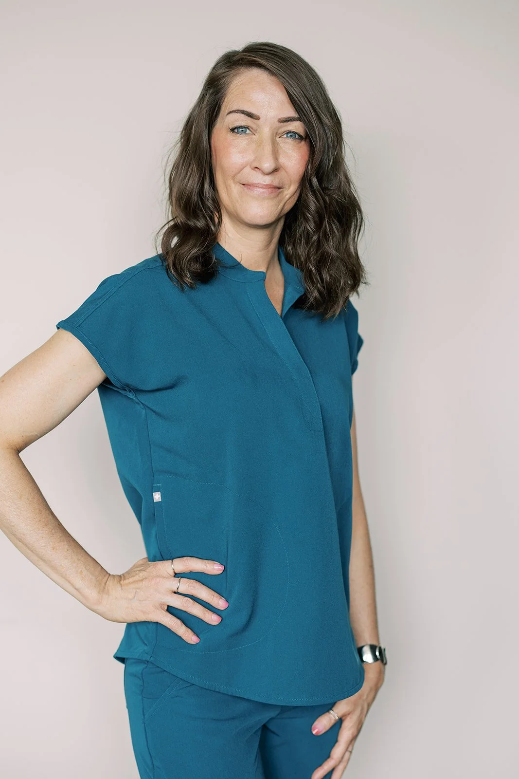 A woman with shoulder-length brown hair, blue eyes, and fair skin wearing teal medical scrubs, standing against a plain light-colored background.