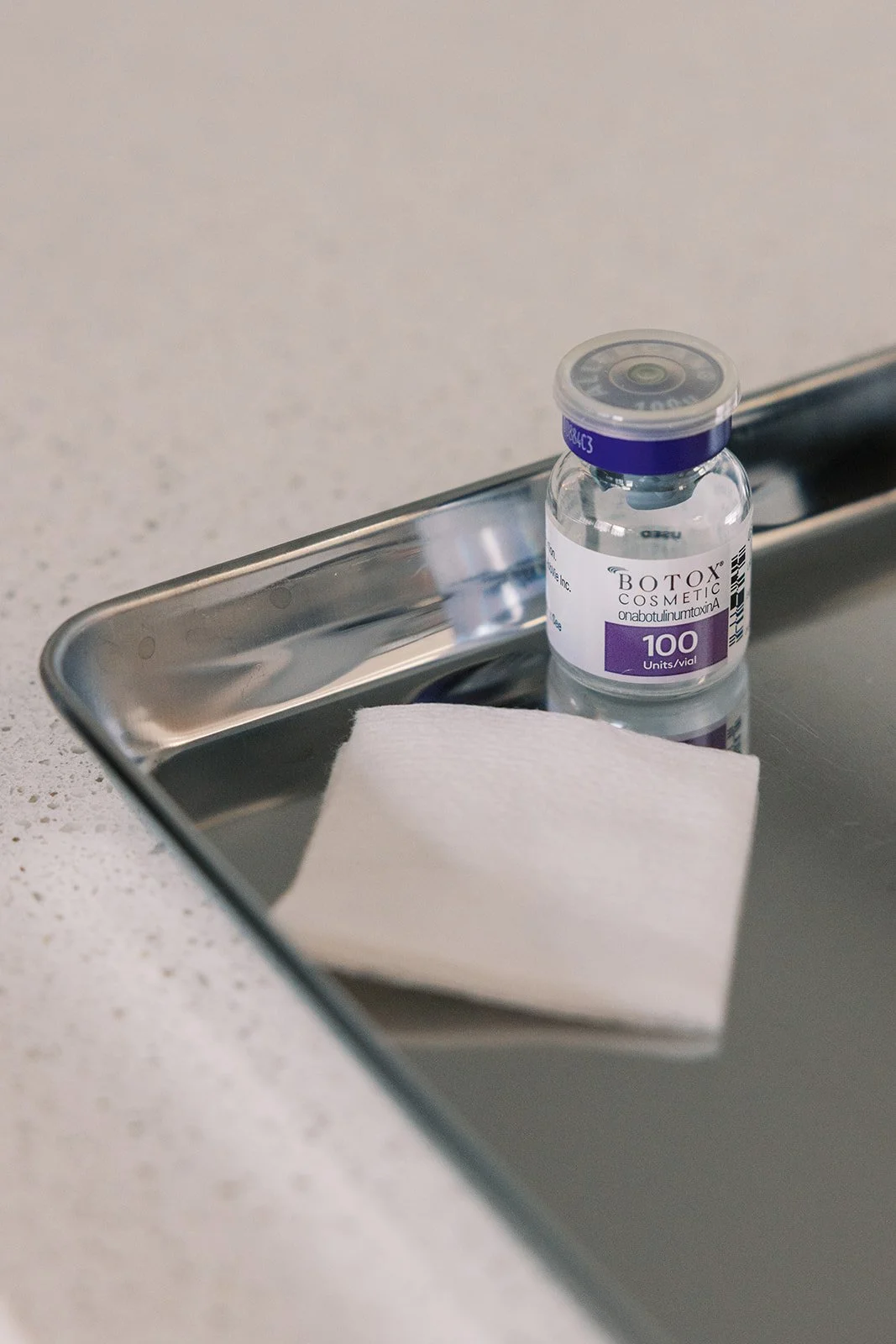 A small glass vial of Botox cosmetic resting on a stainless steel tray with a white paper towel nearby, on a speckled countertop.