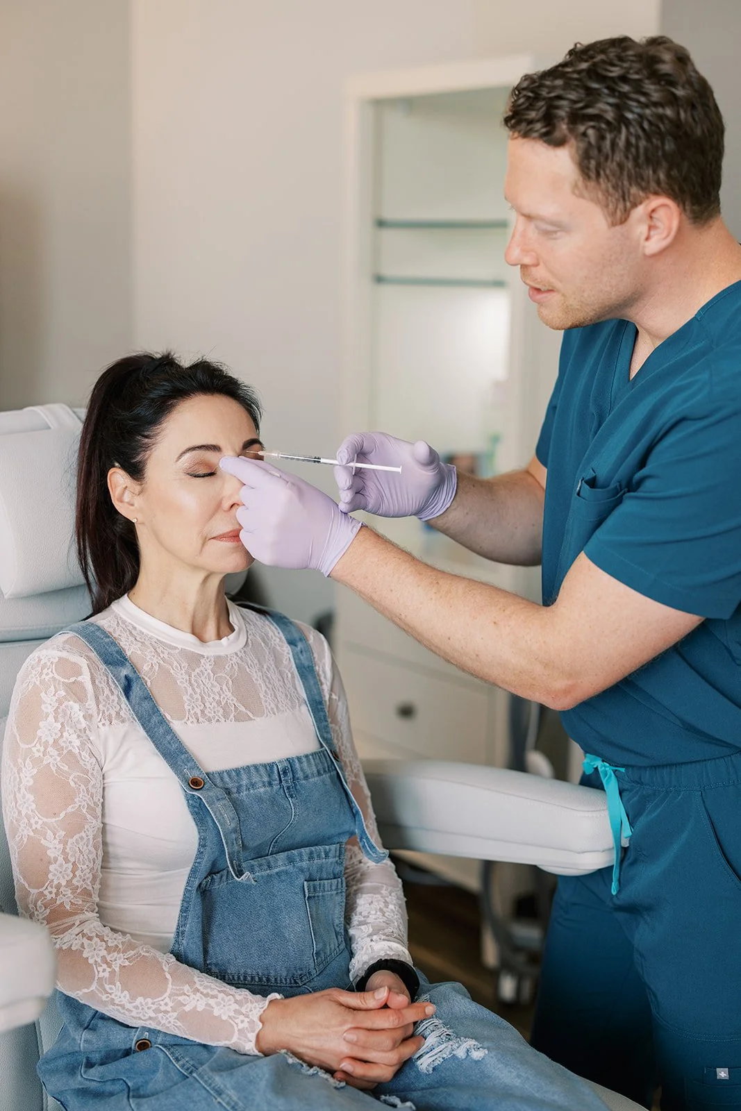 A healthcare professional in scrubs and gloves administering a cosmetic injection to a woman who is sitting and has her eyes closed in a medical setting.
