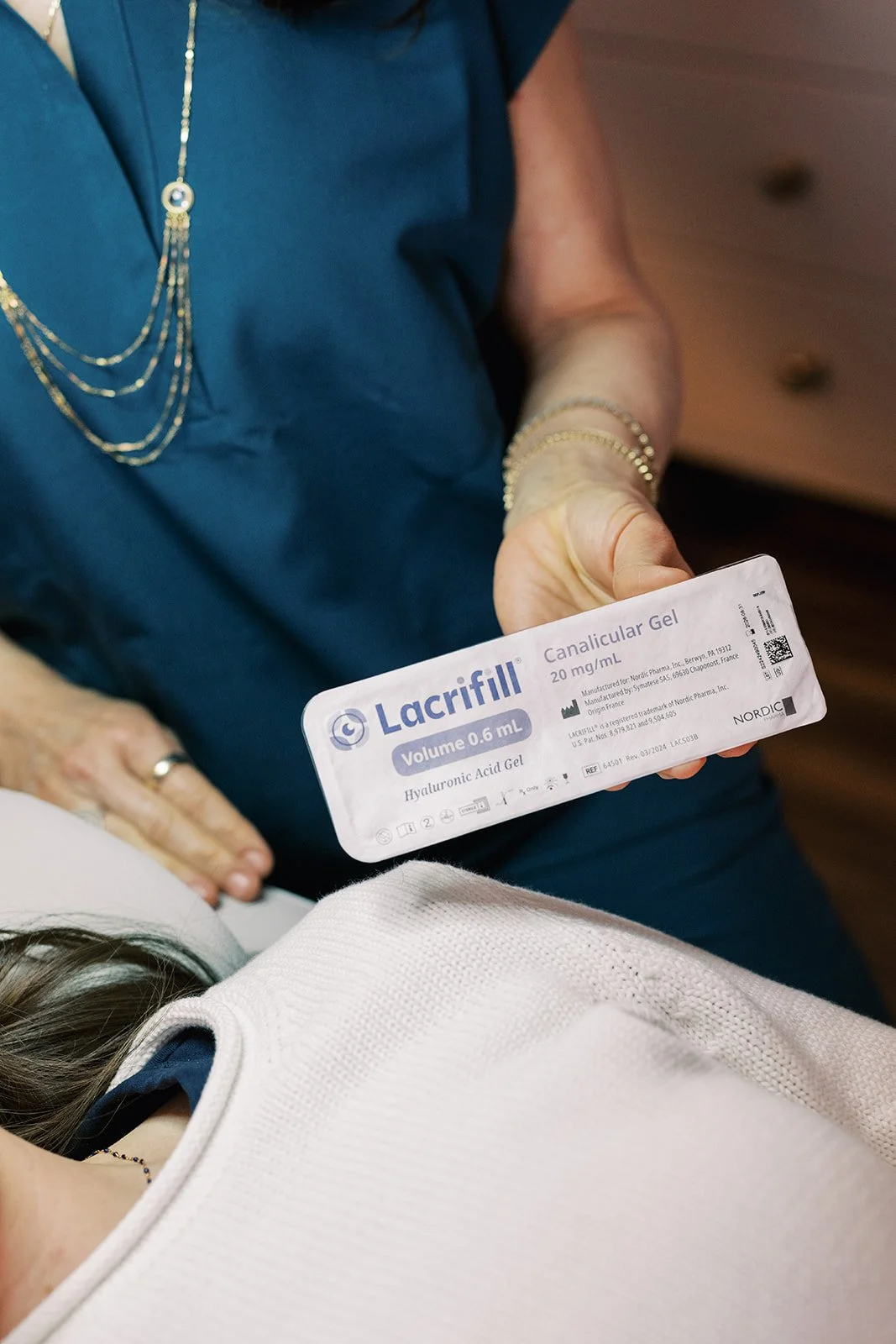 A woman holding a package of Lacrifill hyaluronic acid gel, with a person lying down in front of her. The woman is wearing a blue top and jewelry.