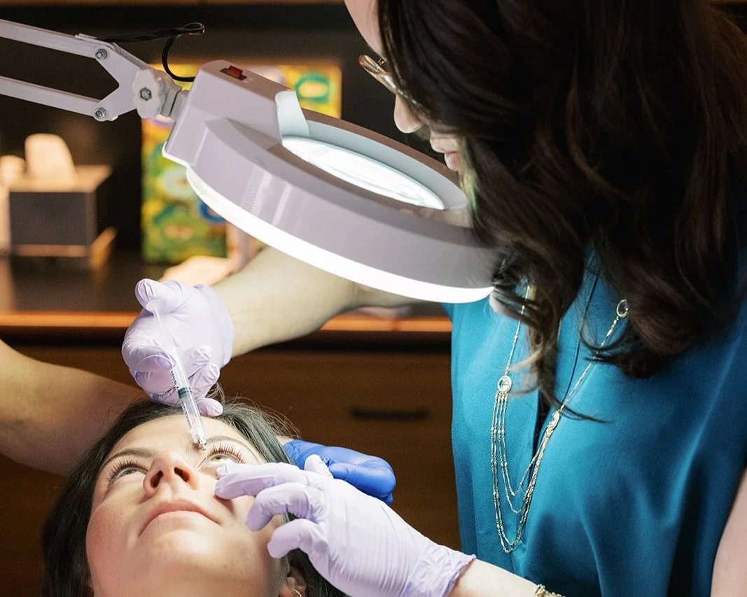A woman receiving a cosmetic or dermatological treatment on her face from a professional in a clinical setting. The practitioner is using a syringe near the woman's at her nose and is wearing gloves. A magnifying lamp is positioned above them for detailed work.