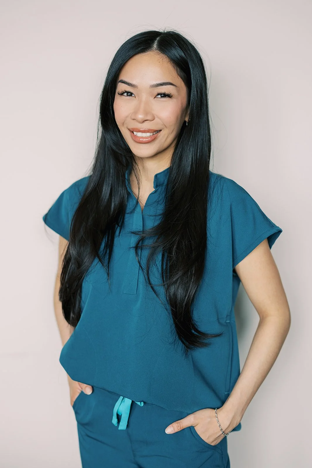 Woman with long black hair smiling and wearing blue scrubs, posed against a plain light-colored background.