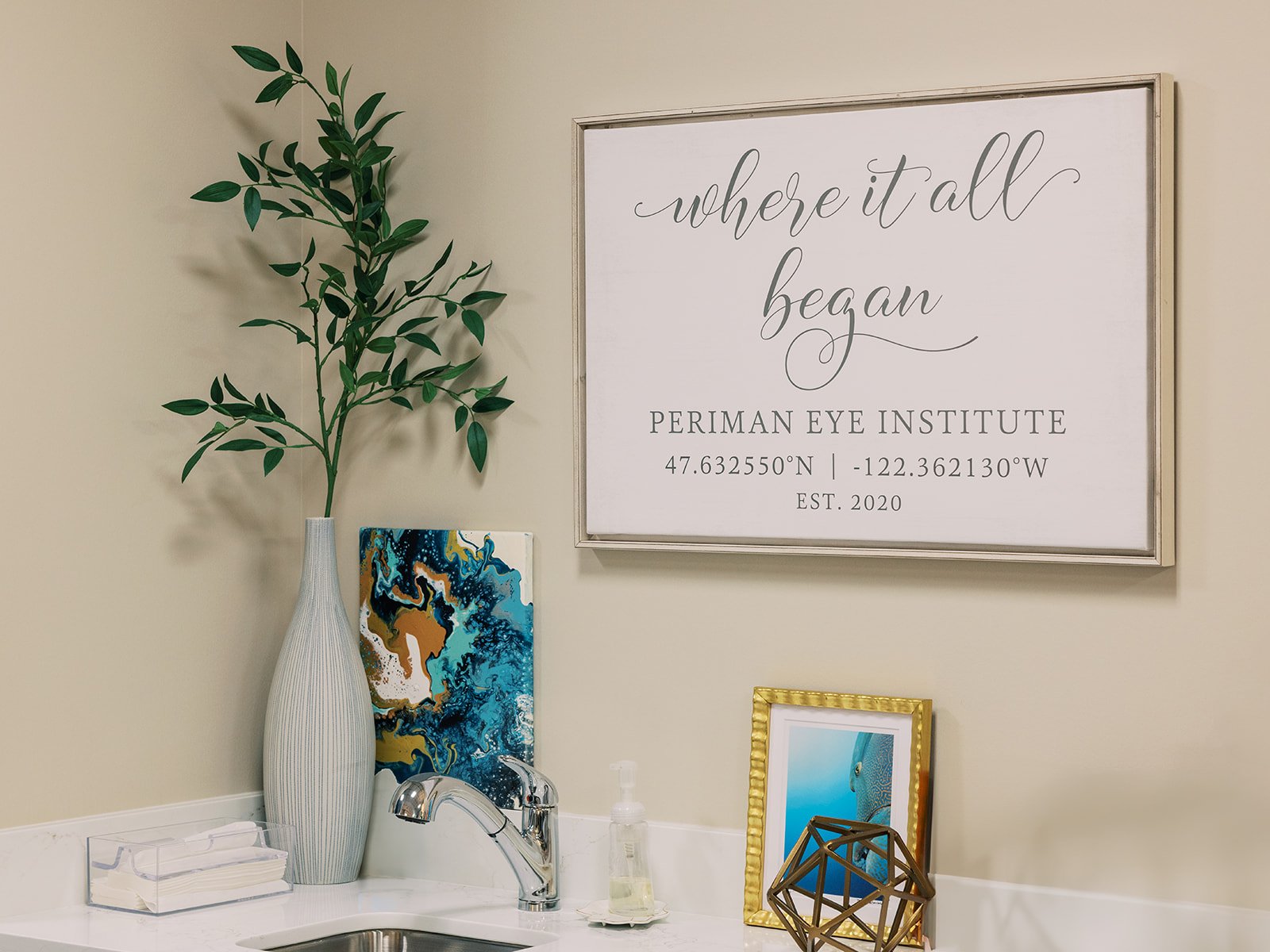 A wall with a framed sign that reads 'where it all began' at the Periman Eye Institute, with coordinates and EST 2020. Below, a white vase with green leafy branches, a colorful abstract painting, a framed photograph, and decorative objects on a white countertop.