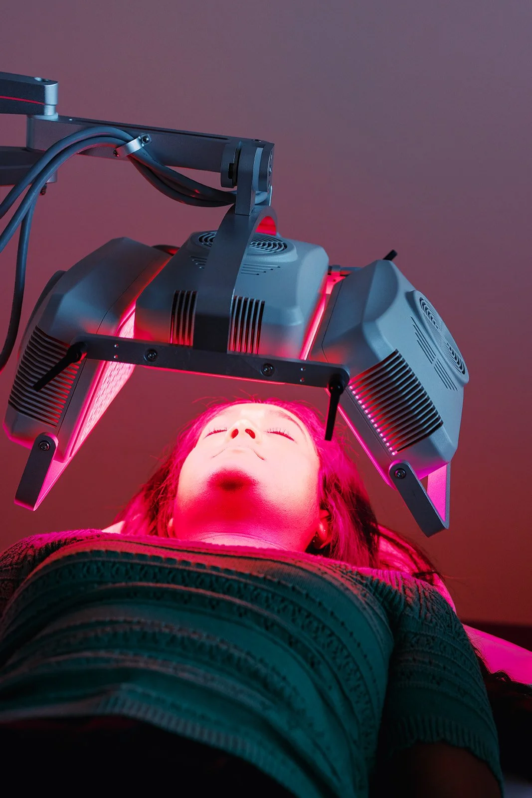 Woman lying down with eyes closed under pink-red LED light therapy device.
