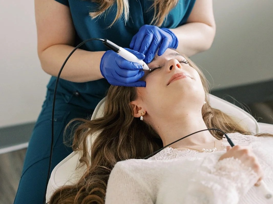 A woman lying down receiving a cosmetic facial treatment from a technician wearing blue gloves, using a handheld device.