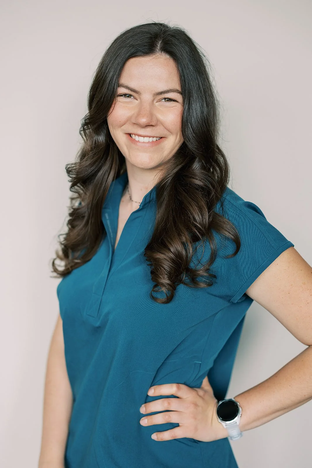 A woman with long, dark, wavy hair smiling and wearing a blue short-sleeved shirt, standing against a plain light background, with her left hand on her hip and wearing a smartwatch.