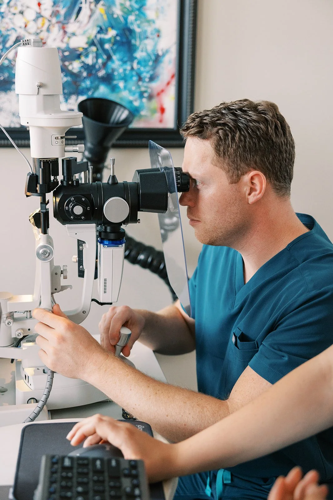 Male eye doctor or optometrist examining a patient's eye using a slit lamp microscope in a clinical setting.