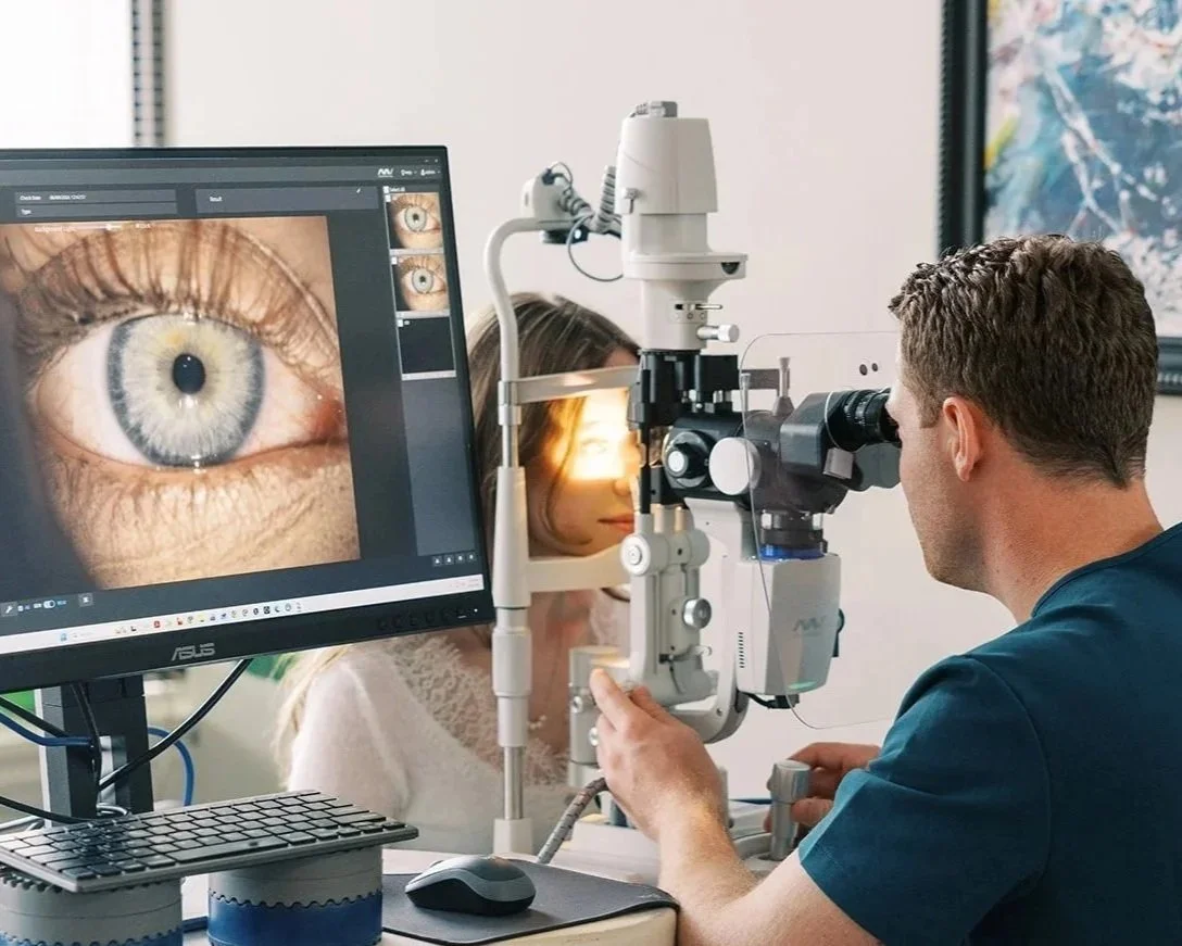 Optometrist examining a woman's eye using specialized equipment in an eye clinic, with a close-up of an eye displayed on the computer monitor.