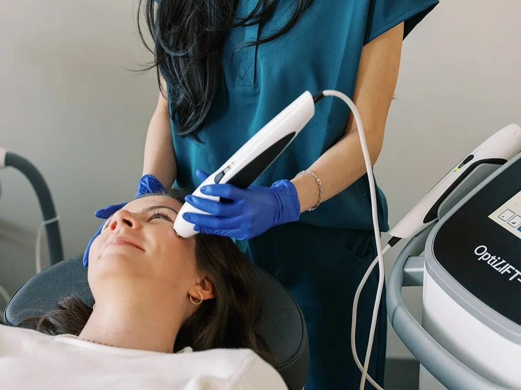 A woman is receiving a cosmetic facial treatment from a technician using a handheld device, in a clinical setting.