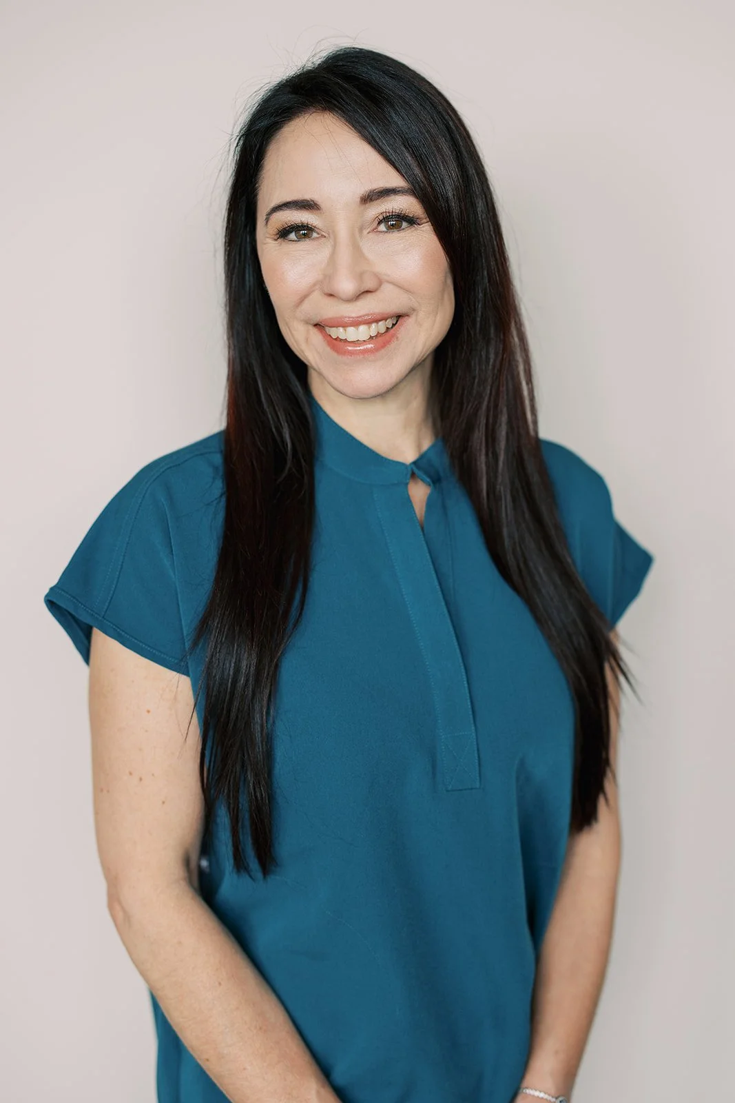 A woman with long dark hair wearing a teal blouse, smiling with teeth showing, against a plain light background.