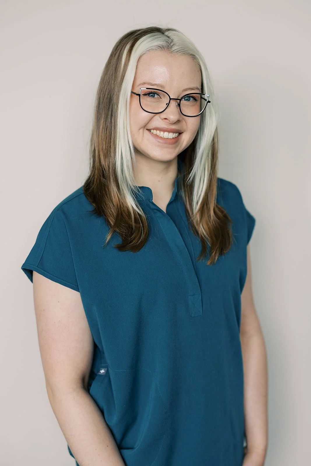 A woman with long, partly blonde and brown hair, wearing glasses and a blue medical uniform, smiling against a plain light-colored background.