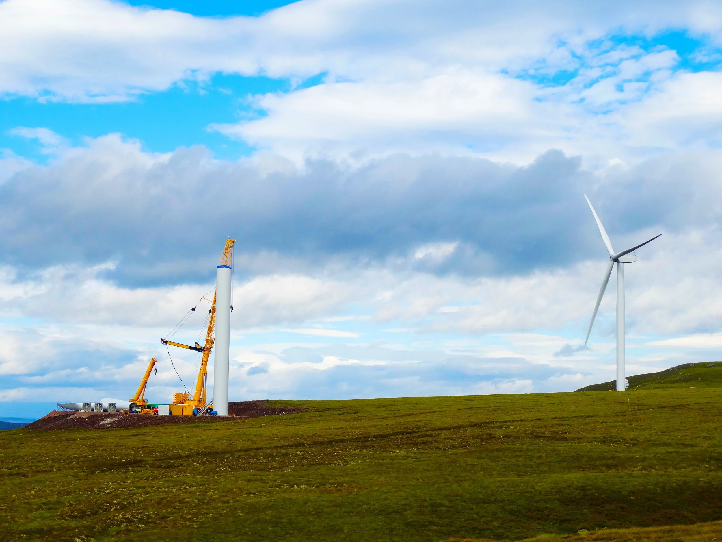 A wind farm under a partly cloudy sky, with two wind turbines and a yellow crane on the ground.