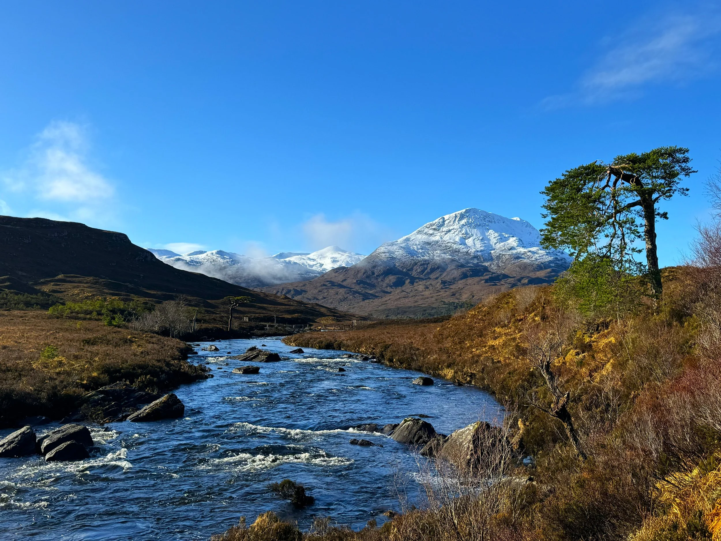 A river flowing through a valley with mountains in the background, snow-covered peaks, and a clear blue sky.
