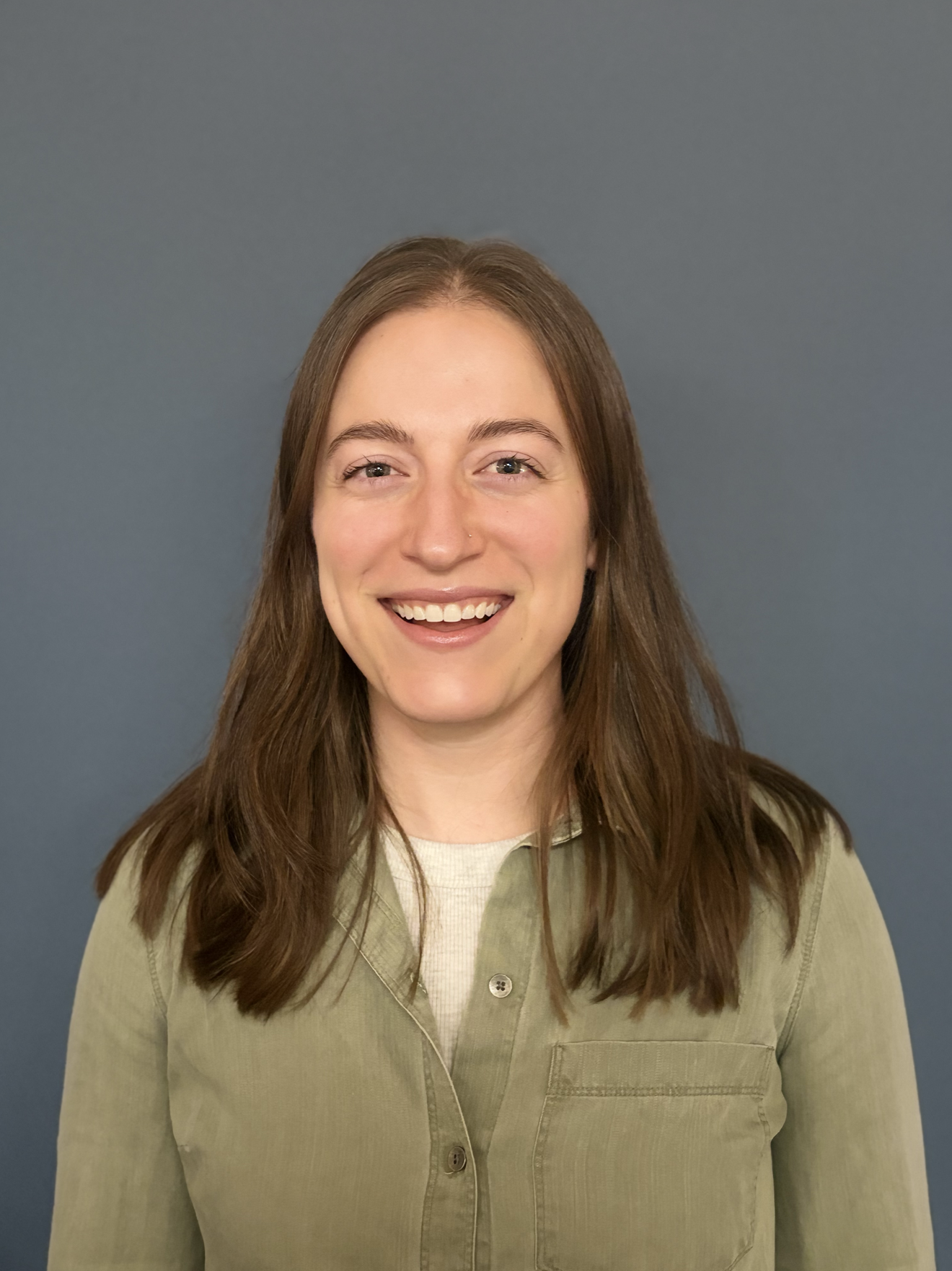 A woman with brown hair smiling, wearing a beige button-up shirt, standing in front of a plain blue wall.