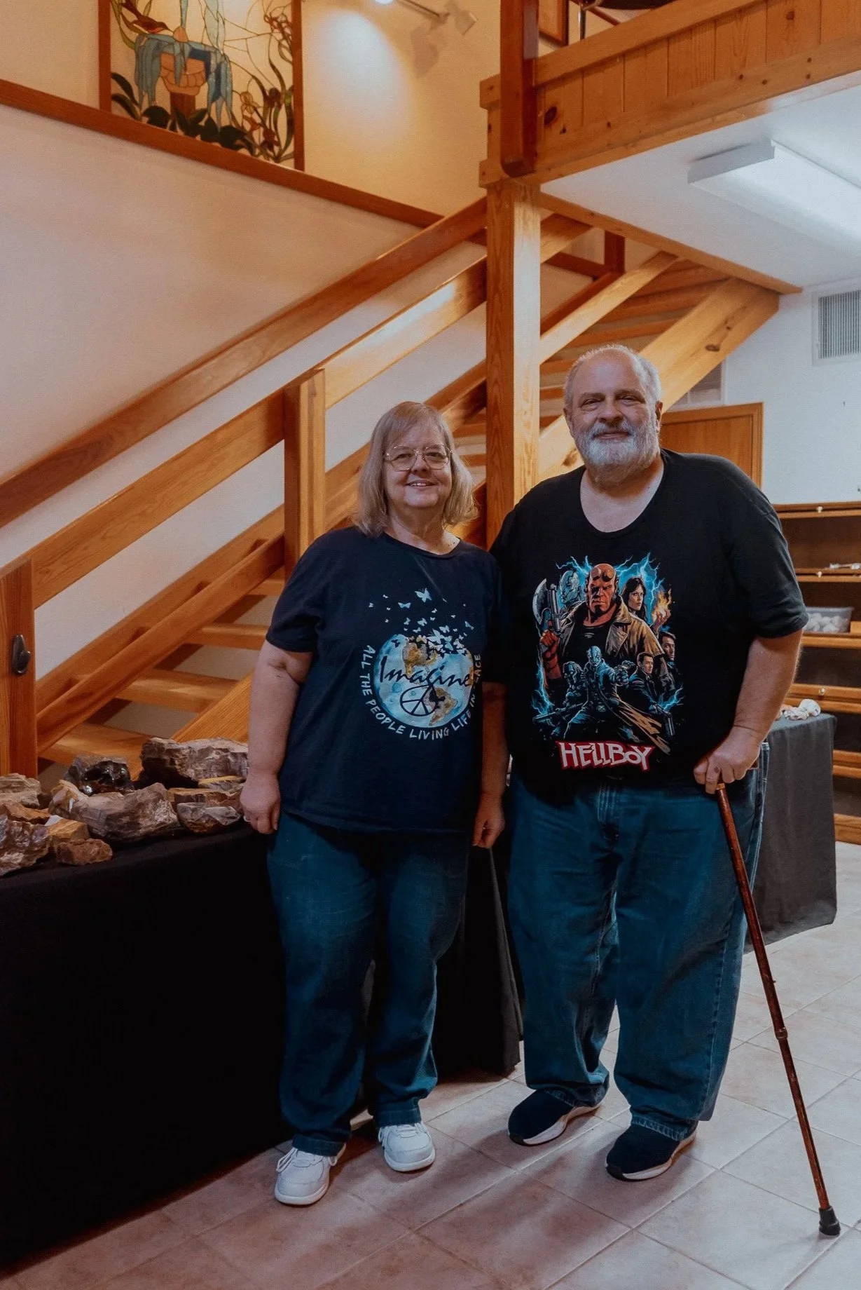 Evil Ed and Kelly standing indoors near a staircase with wooden railings. The woman is wearing a black t-shirt with a graphic design and blue jeans, and the man is wearing a black t-shirt and jeans. Rocks are displayed on a table beside them.