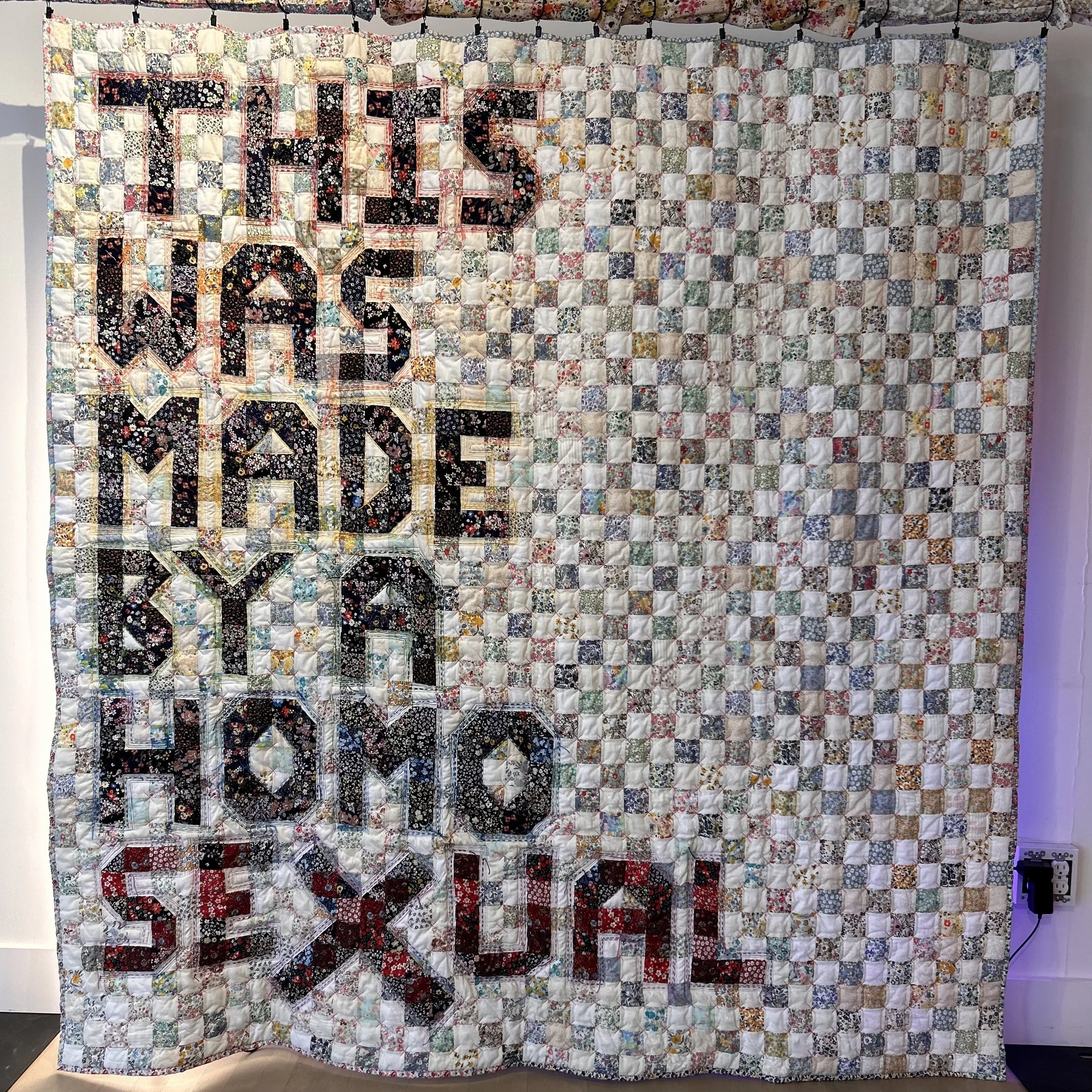 A large quilt hanging on a wall, featuring a patchwork of small floral fabric squares and a large central design with the word 'LOVE' in black fabric with a floral pattern.