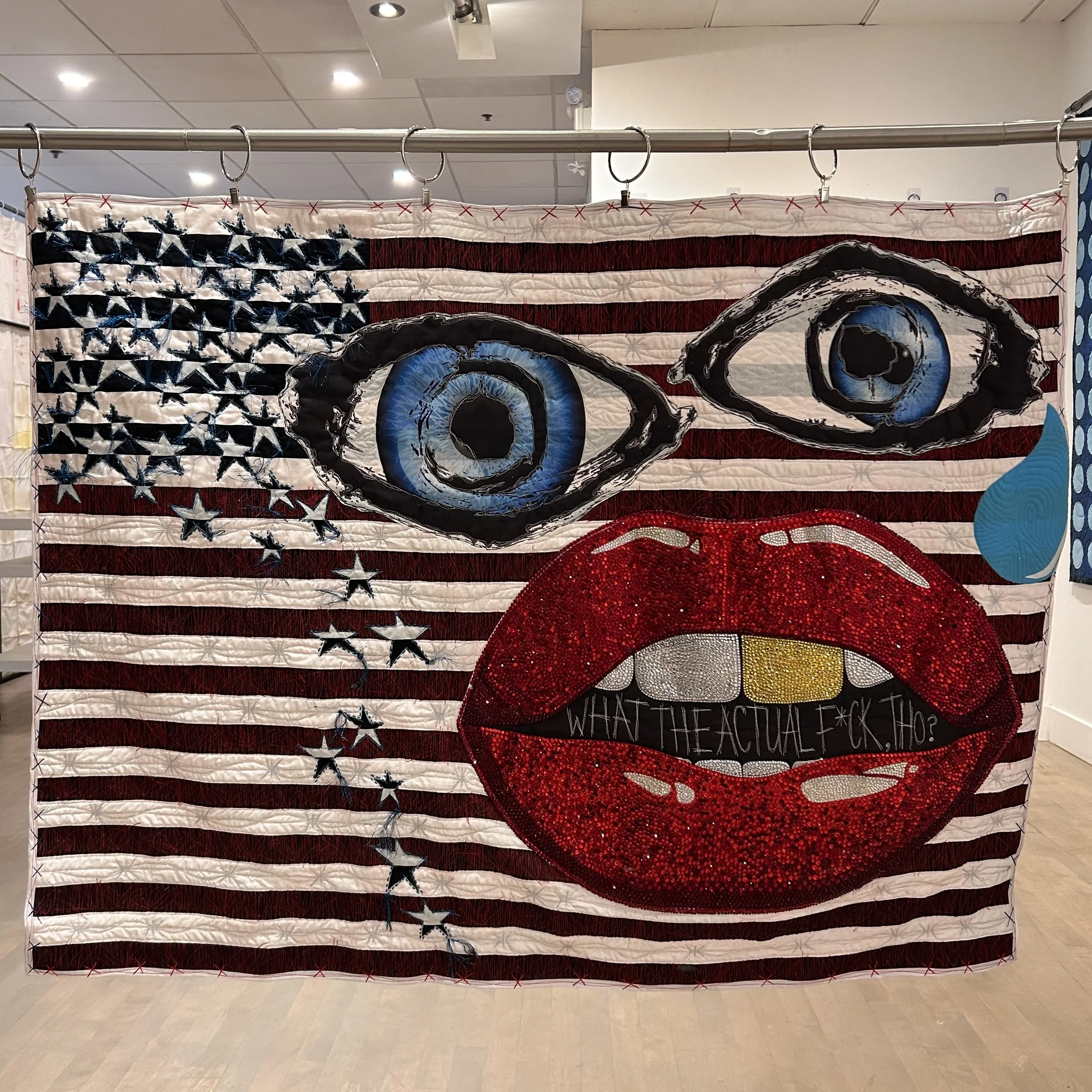 A quilted fabric artwork resembling the American flag with large eyes and a mouth, incorporating stars and stripes and a red, shiny, textured area at the bottom. A sign below reads: 'WHAT THE ACTUAL F***?'